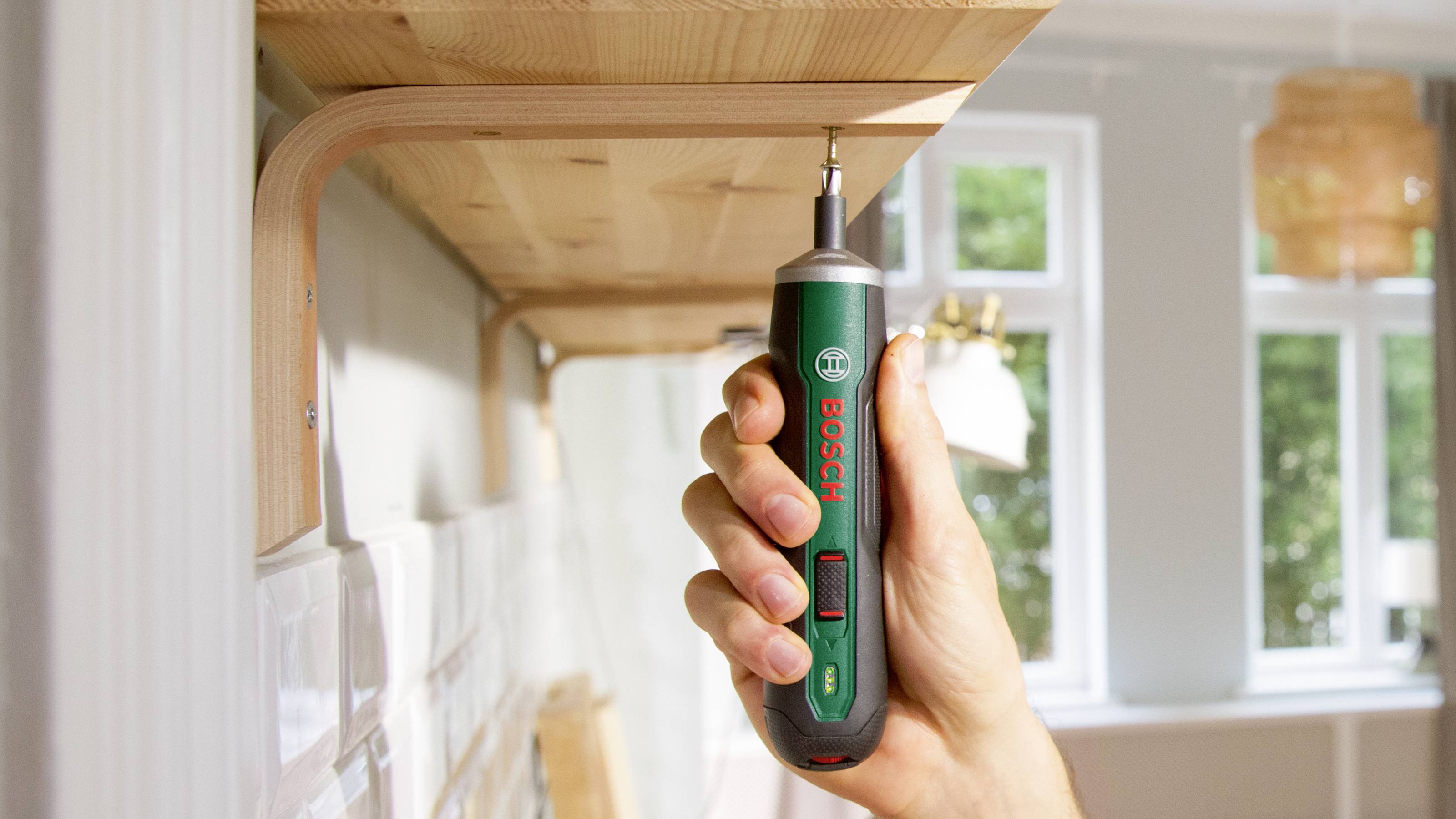 Eine Person hält einen Elektro-Schraubenzieher bei der Montage eines Holzregals. Hintergrund mit unscharfem Raum und Fenstern.