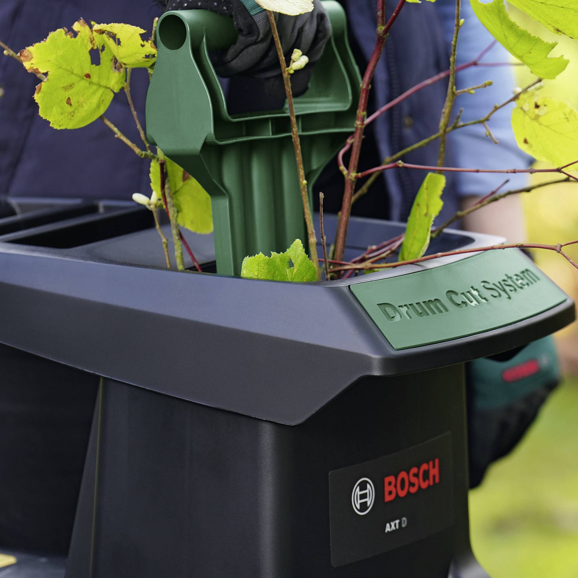 Nahaufnahme einer schwarzen Gartengerätstation mit der Aufschrift 'Drum Cut System' und Bosch-Logo. Ein grünes Gartengerät mit Pflanzen darauf.