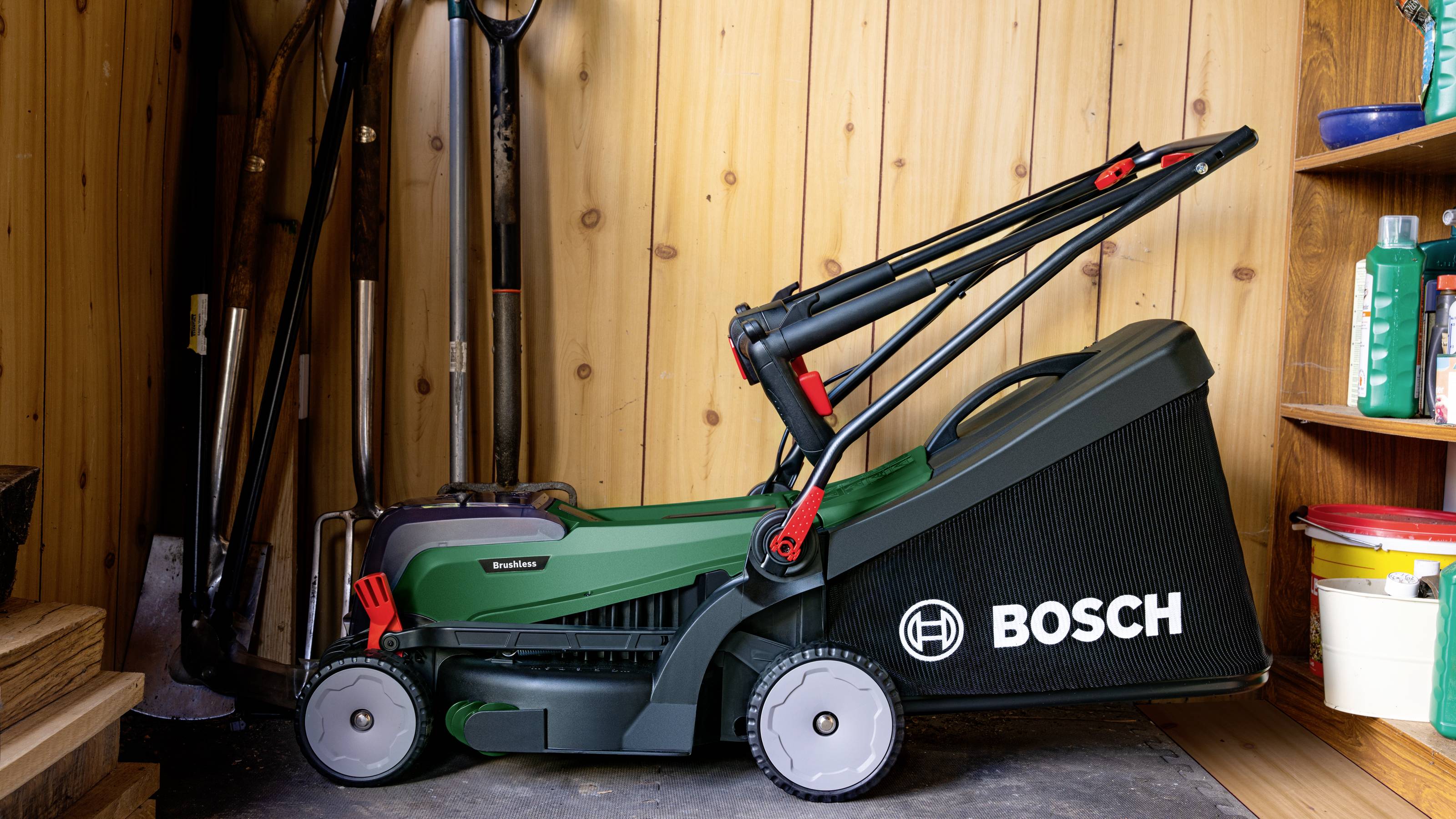 Ein Rasenmäher der Marke Bosch steht in einem hölzernen Schuppen, umgeben von Gartenwerkzeugen und Vorratsregalen.