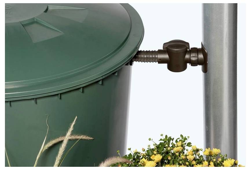 Grüne Mülltonne, die an einem Metallpfosten befestigt ist, mit Blumen am unteren Ende vor einem einfachen, hellen Hintergrund.