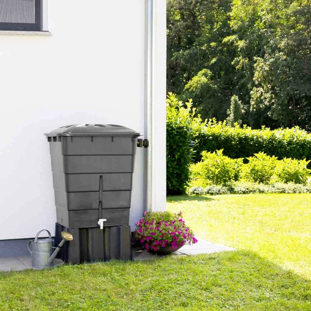 Ein Regenwassertank mit Zapfhahn steht neben einem Haus, umgeben von einem Garten mit grünem Rasen und blühenden Pflanzen.
