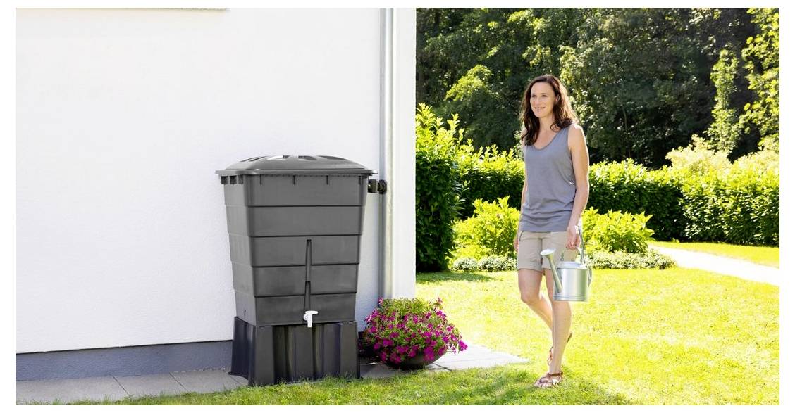 Frau mit einer Gießkanne geht neben einer Regentonne an einem Haus vorbei, mit einem Garten und Blumen im Hintergrund.
