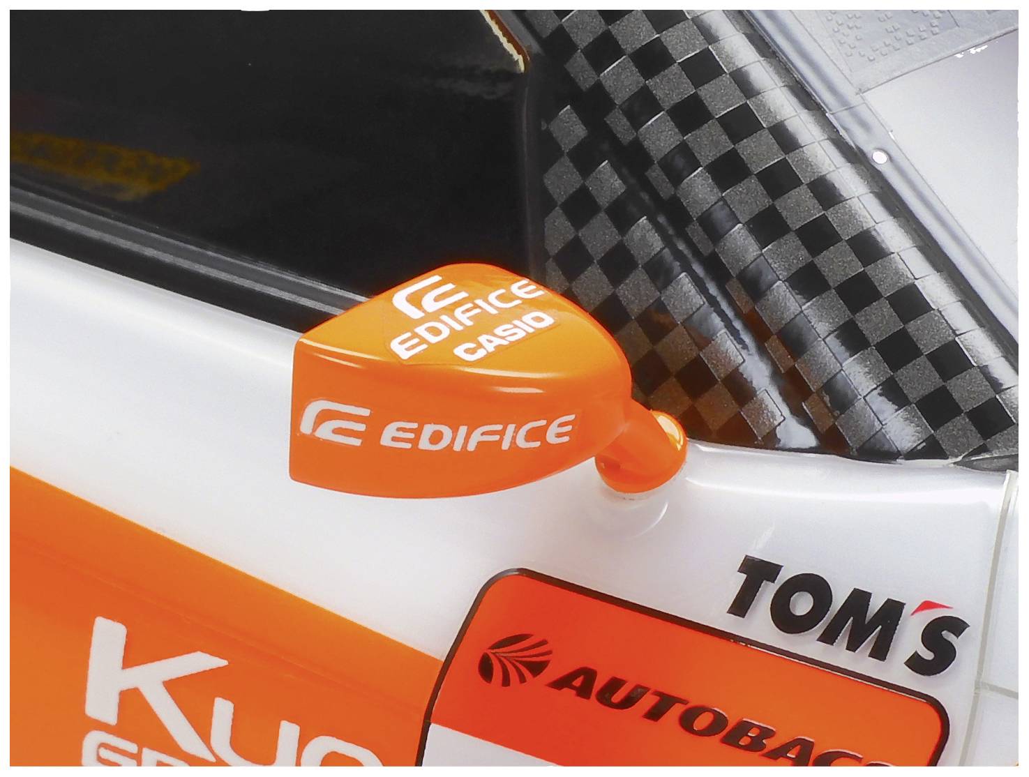 Ein orangefarbener Seitenspiegel eines Rennwagens mit den Logos 'EDIFICE' und 'CASIO', daneben steht 'TOM'S' auf grauem Karomuster.