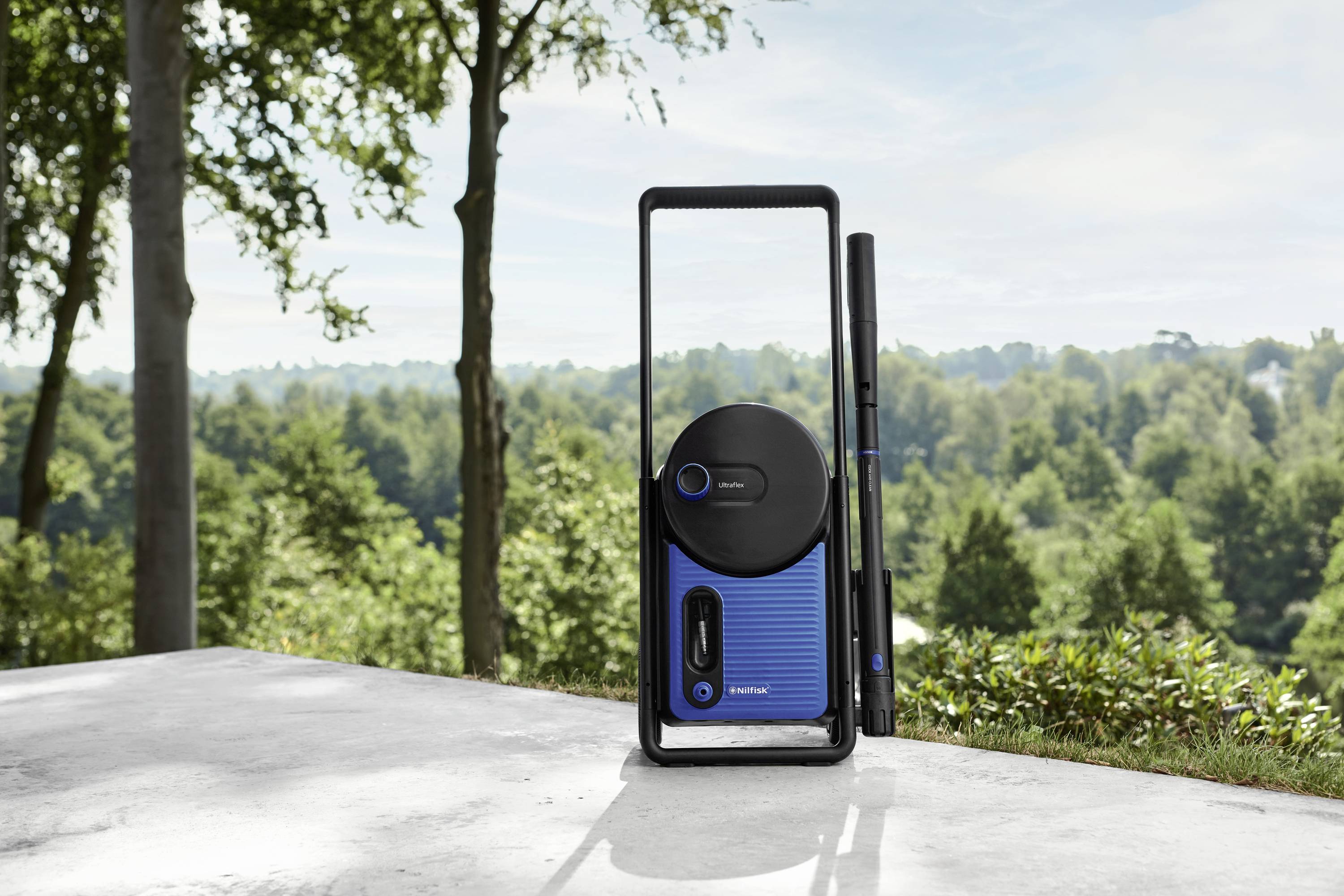 Ein tragbares Lautsprechersystem mit blauem Gehäuse steht auf einer Terrasse mit Blick auf einen bewaldeten Hügel im Hintergrund.