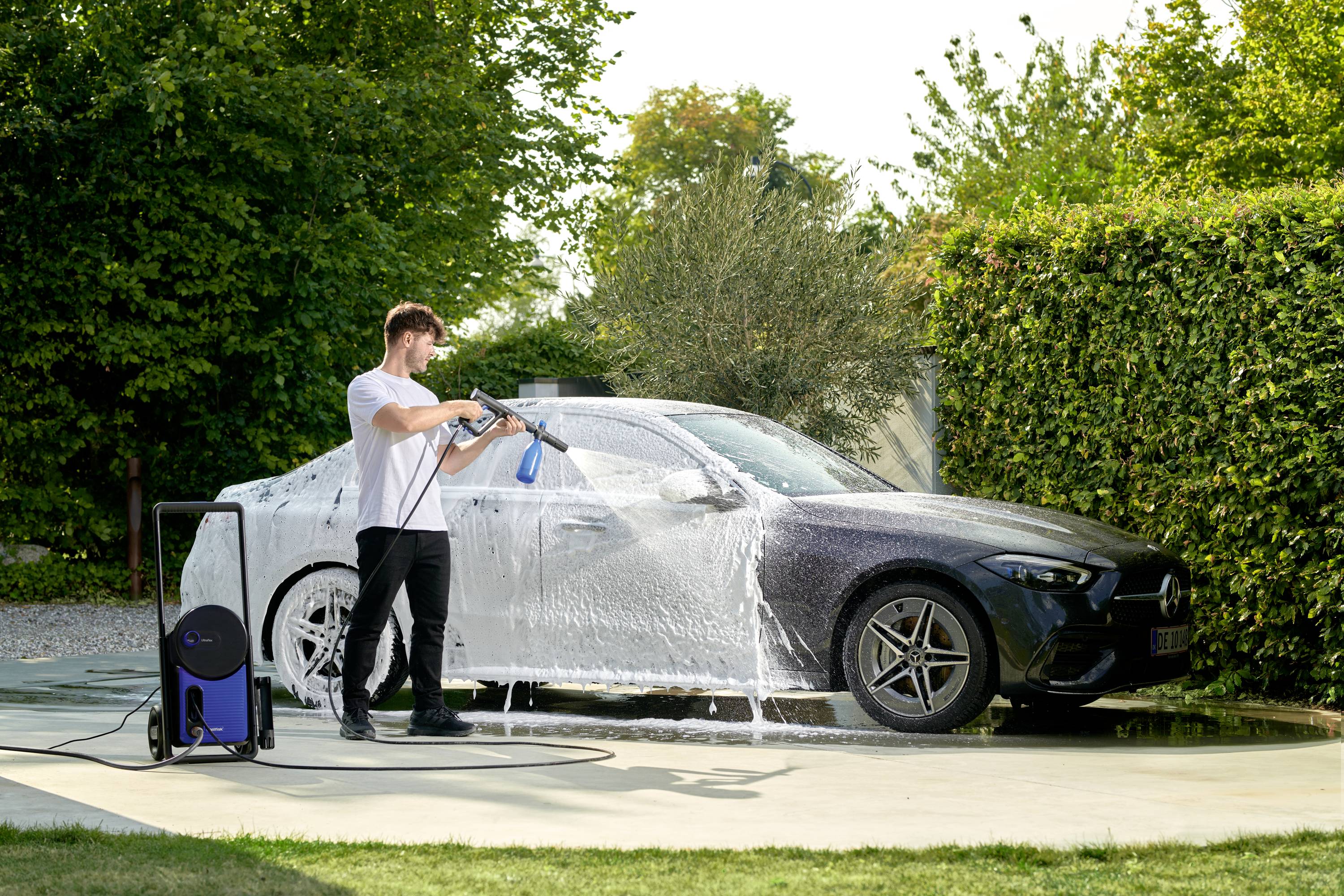 Ein Mann wäscht ein Auto mit einem Hochdruckreiniger in einem Garten. Das Auto ist mit Seifenblasen bedeckt. Im Hintergrund sind Büsche zu sehen.