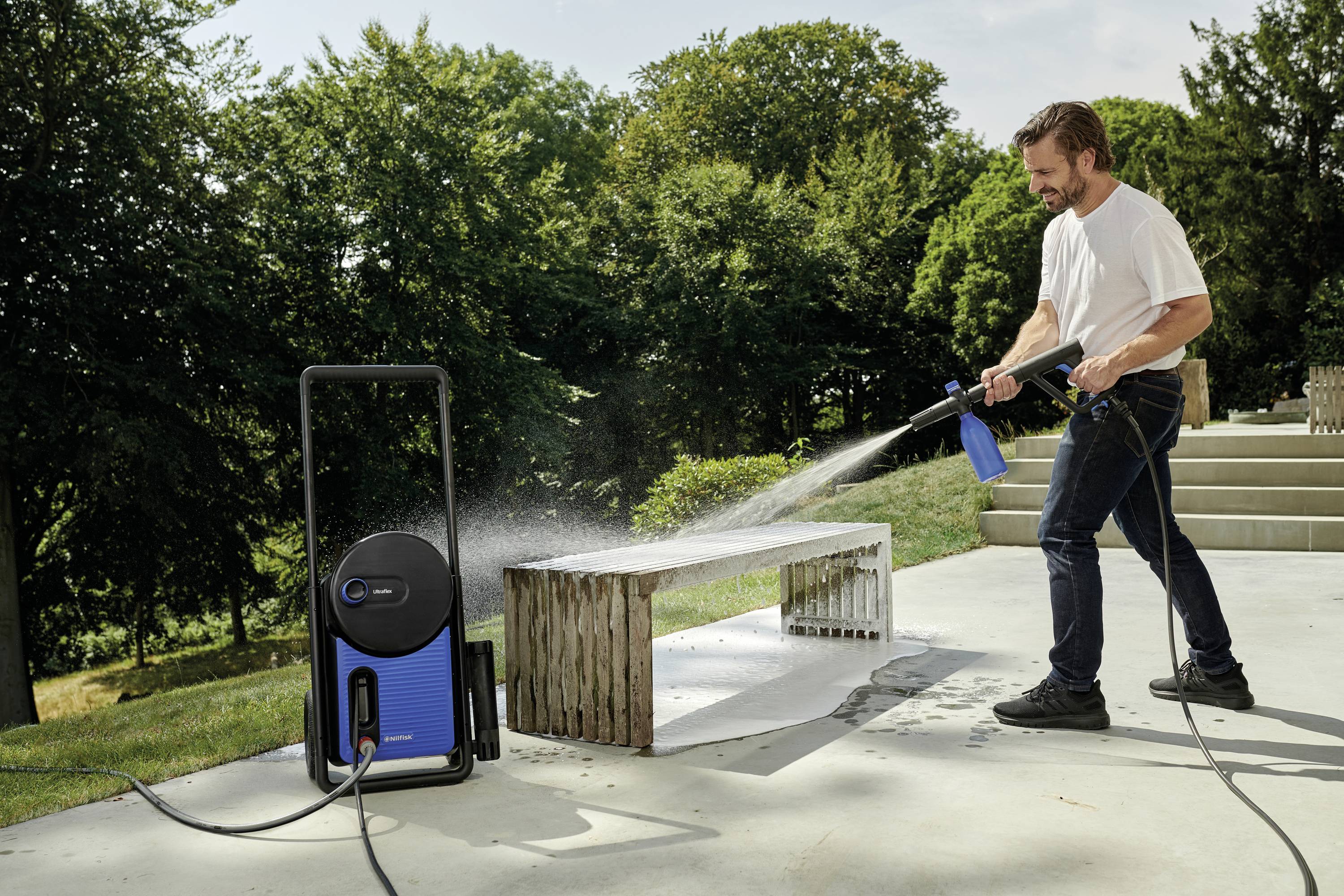 Ein Mann reinigt eine Außenbank mit einem Hochdruckreiniger auf einer Terrasse, umgeben von Bäumen im Hintergrund.