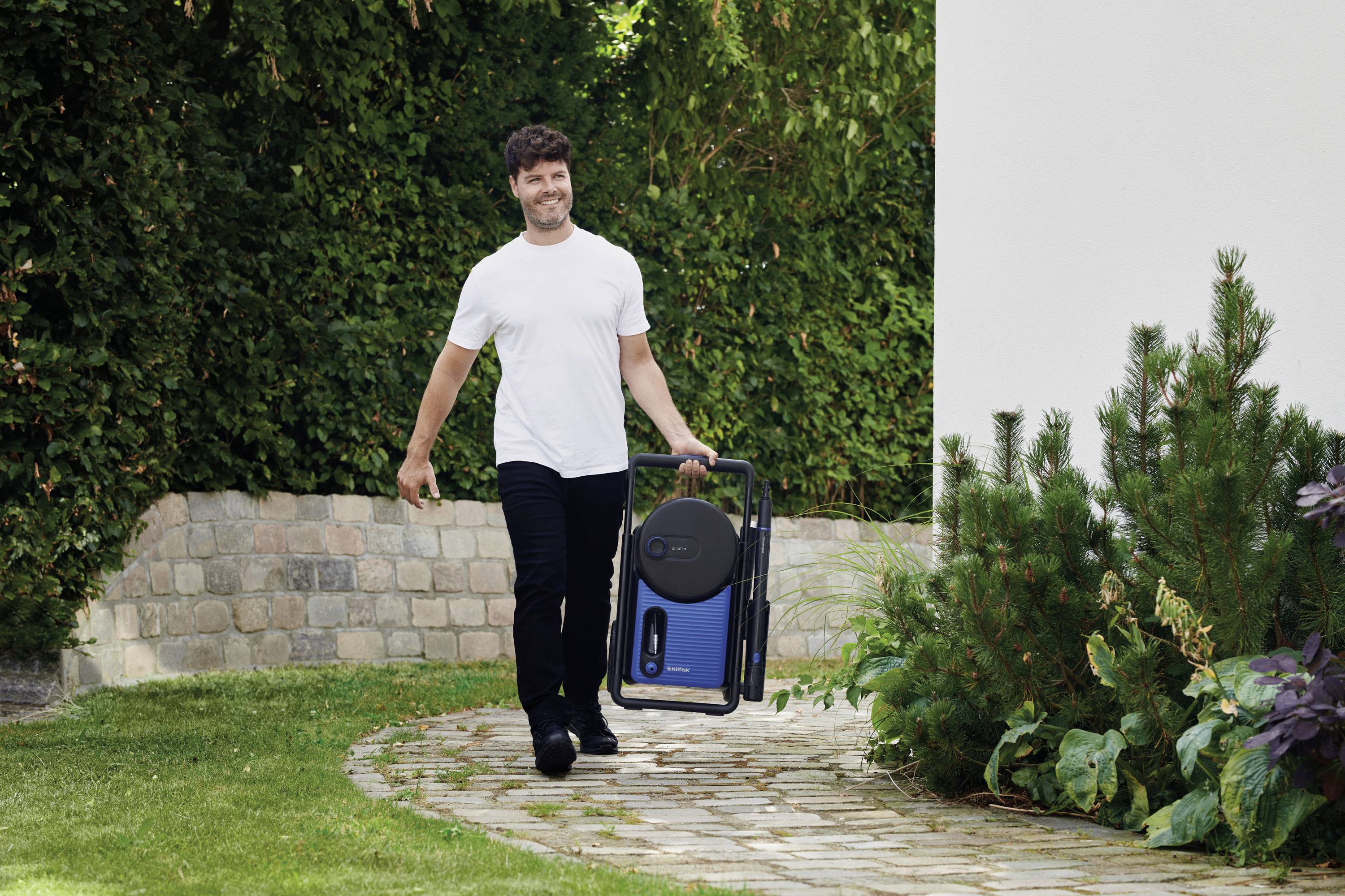Ein Mann geht lächelnd auf einem gepflasterten Gartenweg. Er trägt einen tragbaren, blauen, rechteckigen Gegenstand. Im Hintergrund ist eine dichte grüne Hecke zu sehen.