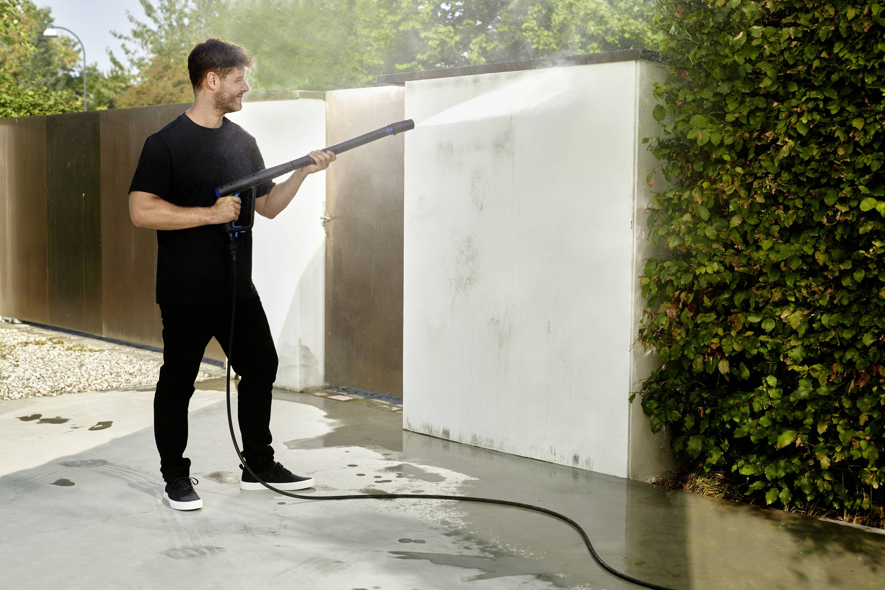 Mann reinigt Wand mit Hochdruckreiniger im Freien. Neutrale Farbgebung, grüne Pflanzen im Hintergrund.