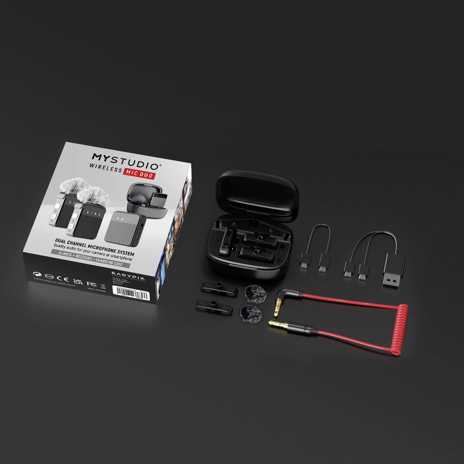 Draufsicht auf MyStudio Wireless Mic Duo: Mikrofone, Ladeetui, Kabel und Zubehör auf schwarzer Oberfläche. Verpackung links.