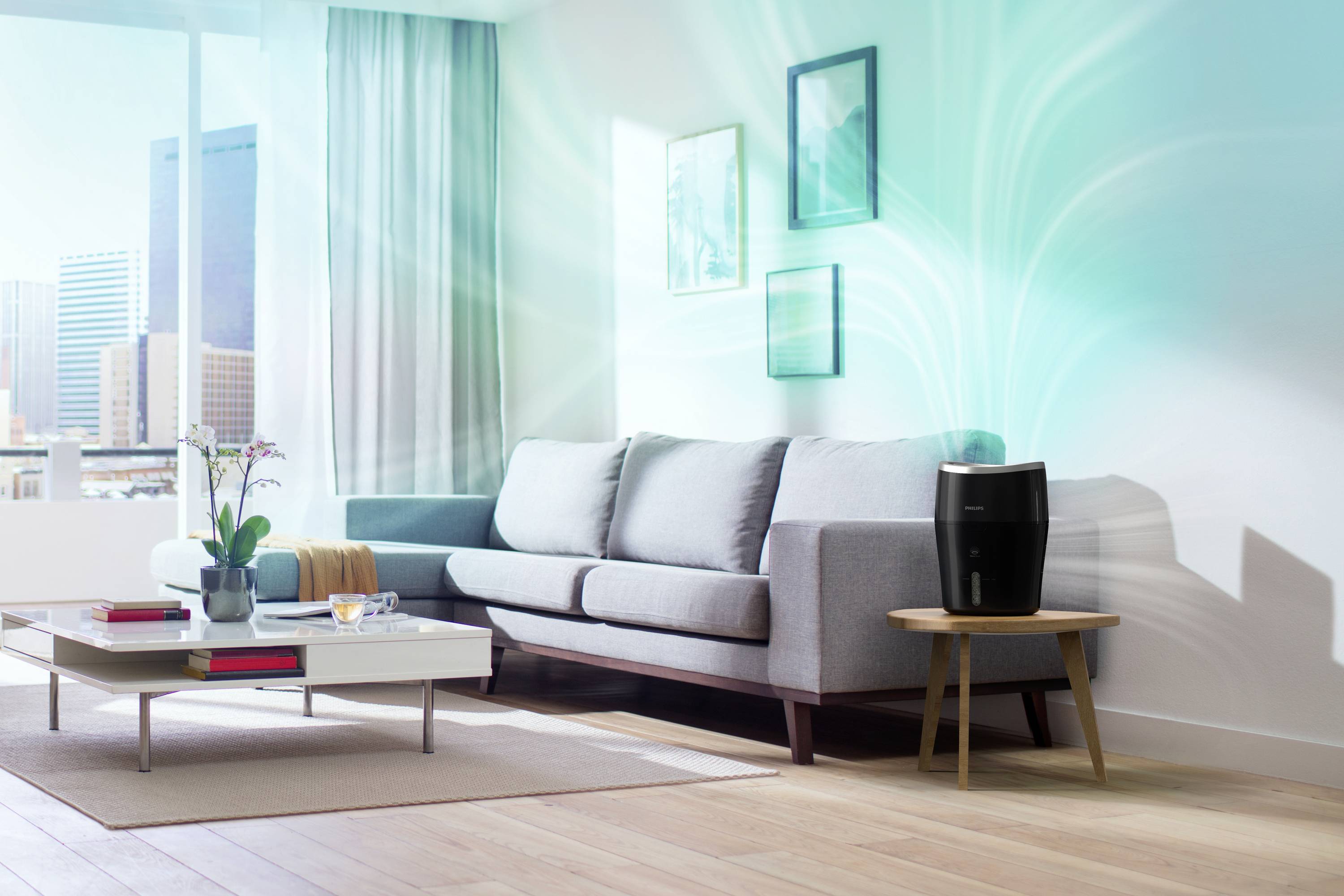 Helles Wohnzimmer mit grauem Sofa, moderner Luftreiniger auf einem Tisch und sichtbare Luftströme, die Frische suggerieren.