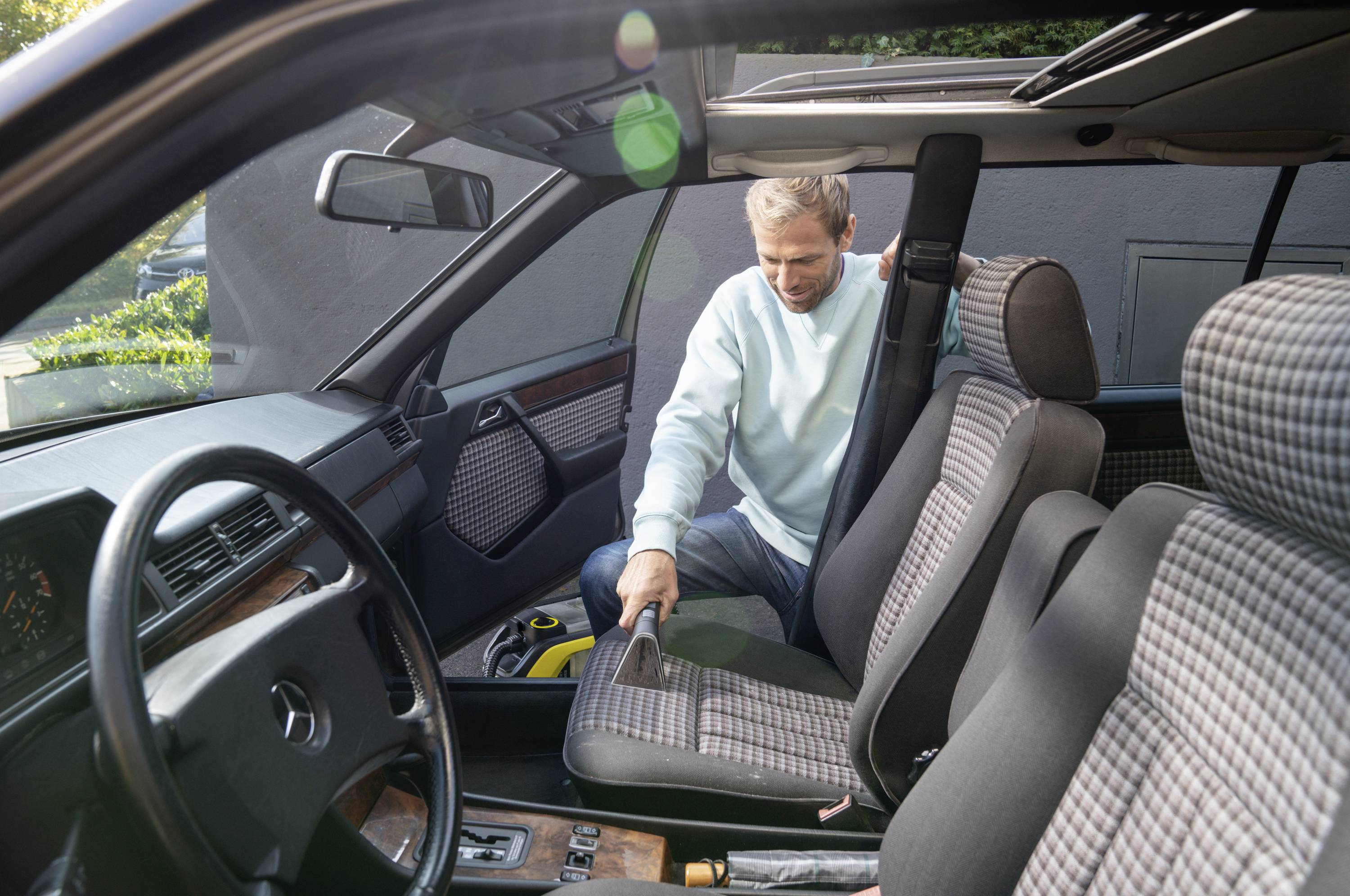 Ein Mann reinigt den Innenraum eines Autos, saugt den Boden. Er trägt einen hellblauen Pullover. Tageslicht fällt durch das Autofenster.