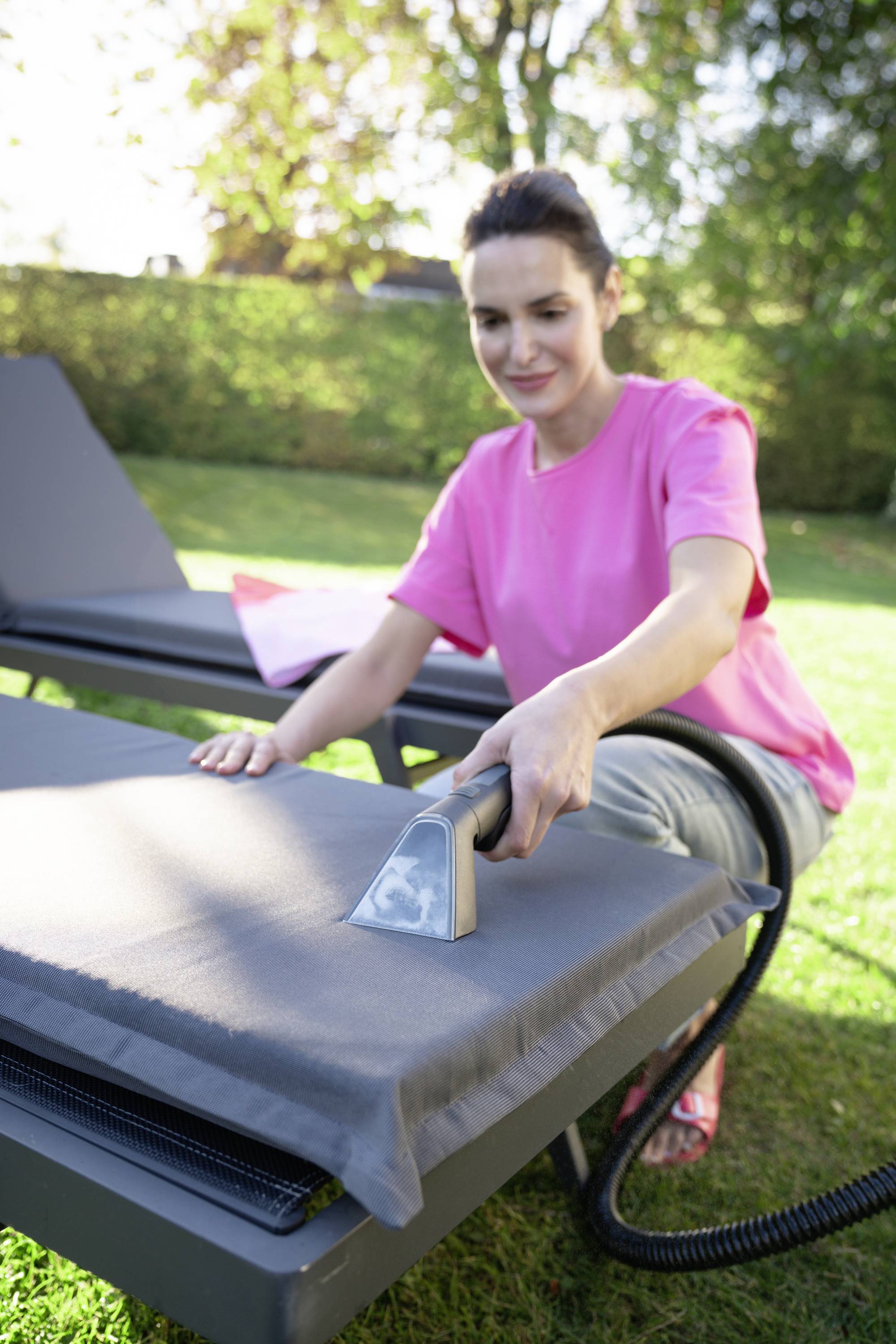 Eine Frau reinigt im Garten mit einem Dampfreiniger das Polster einer Sonnenliege. Im Hintergrund ist grüne Vegetation zu sehen.