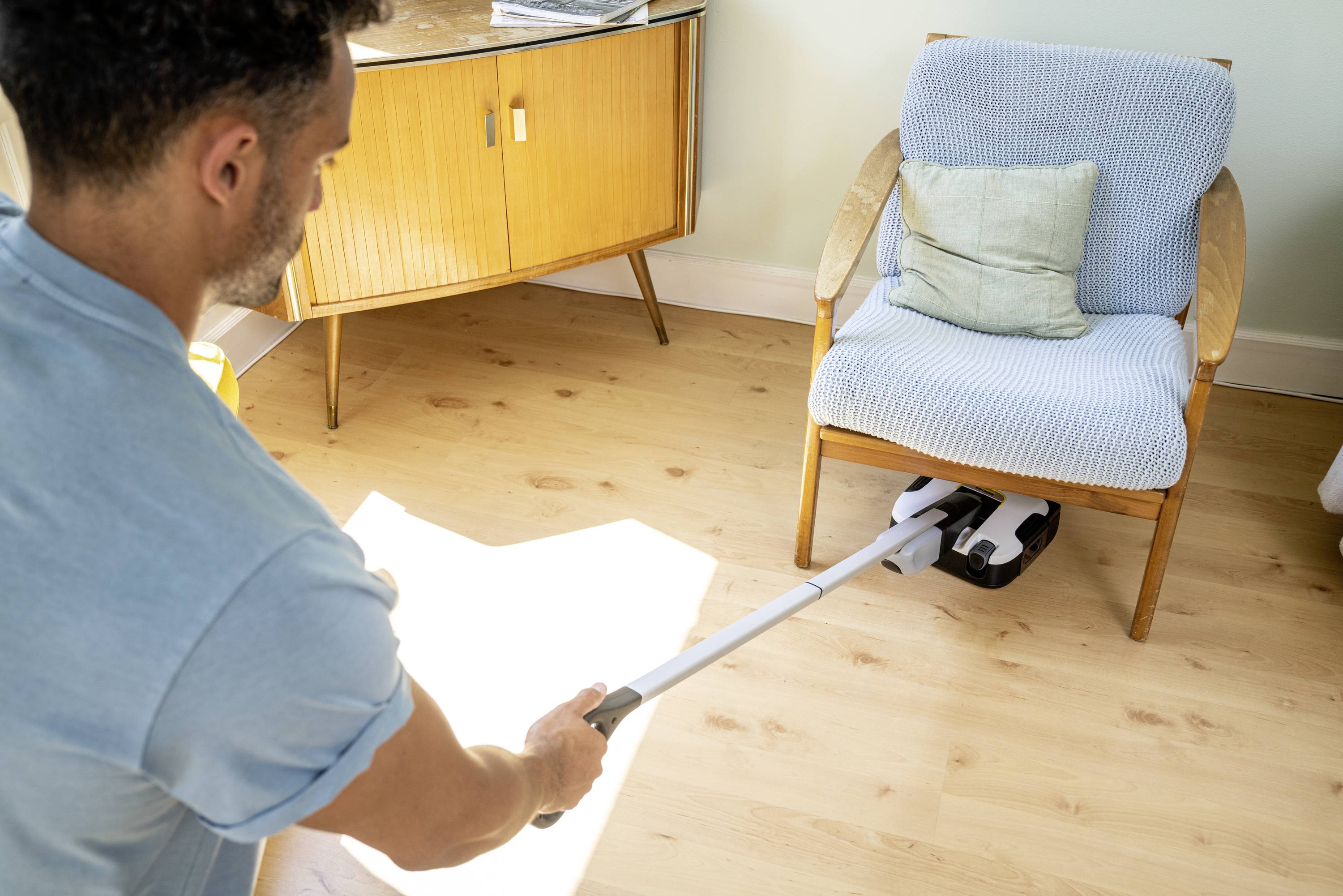Mann saugt Holzfußboden unter einem Sessel in einem hellen Raum. Im Hintergrund steht ein Sideboard.