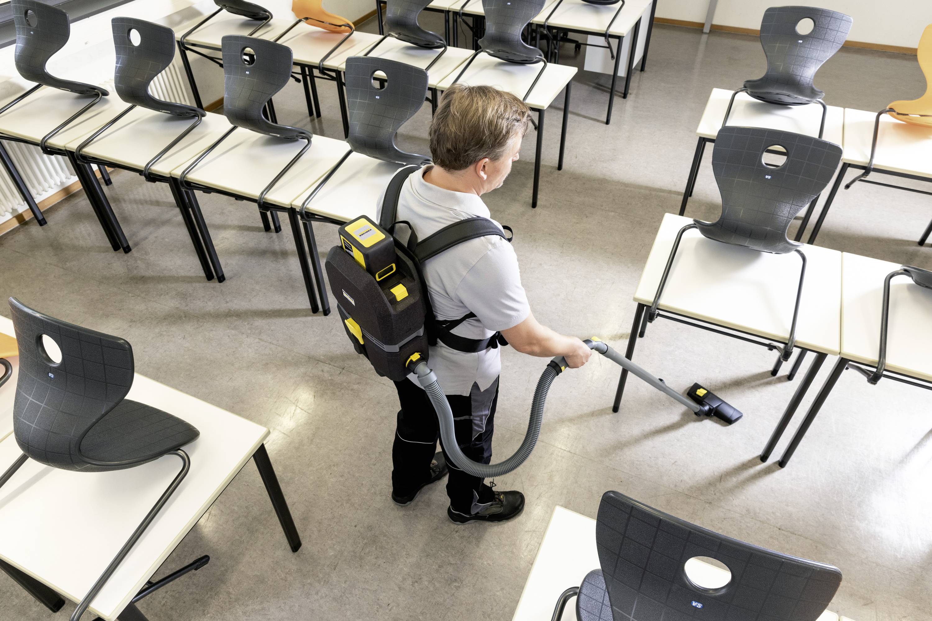 Mann mit Rucksackstaubsauger reinigt einen leeren Klassenraum. Stühle sind hochgestellt, der Boden wird gesaugt.