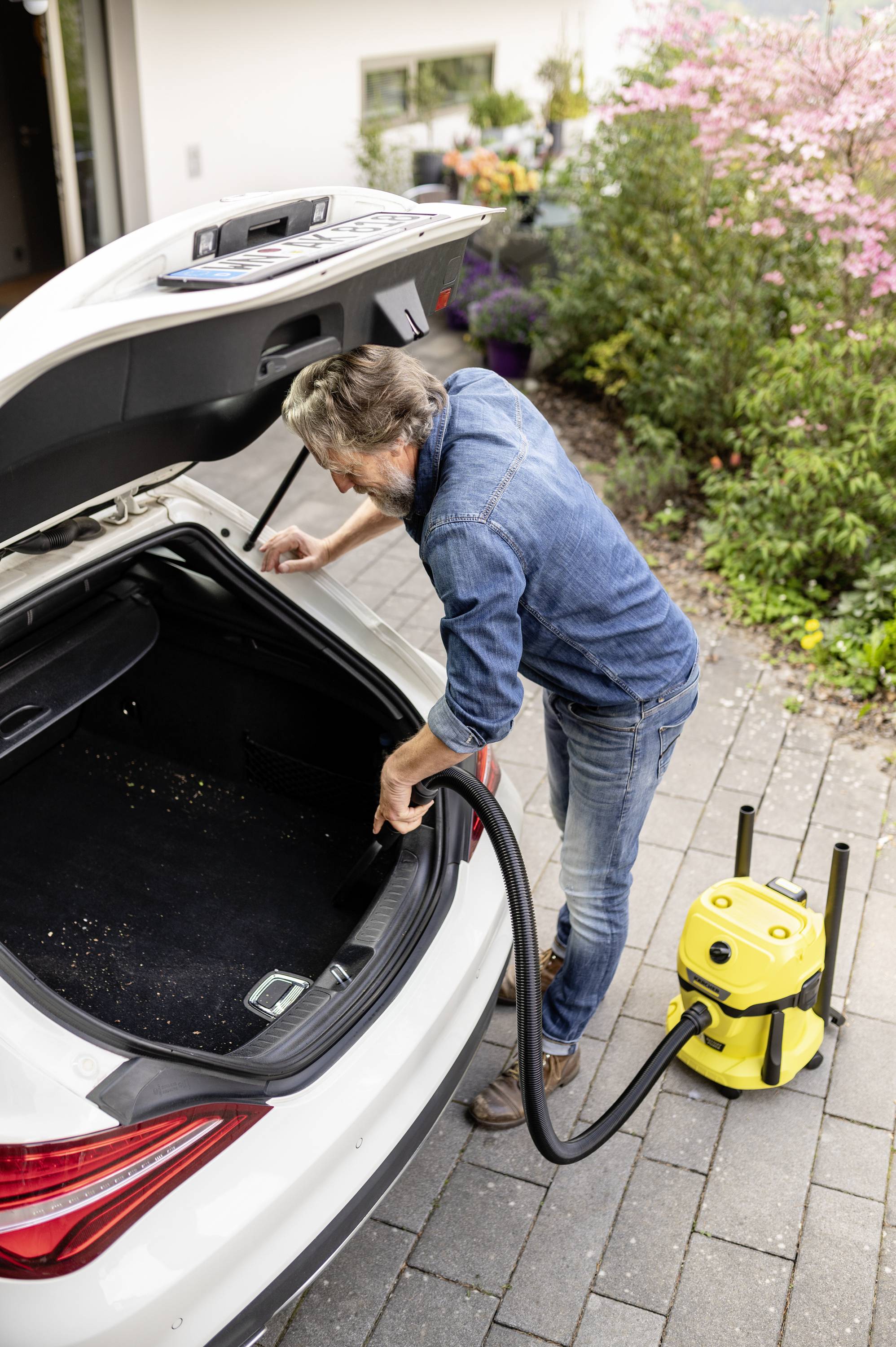 Mann saugt den Kofferraum eines Autos mit einem gelben Staubsauger im Freien. Im Hintergrund Blumen und ein Gebäude.