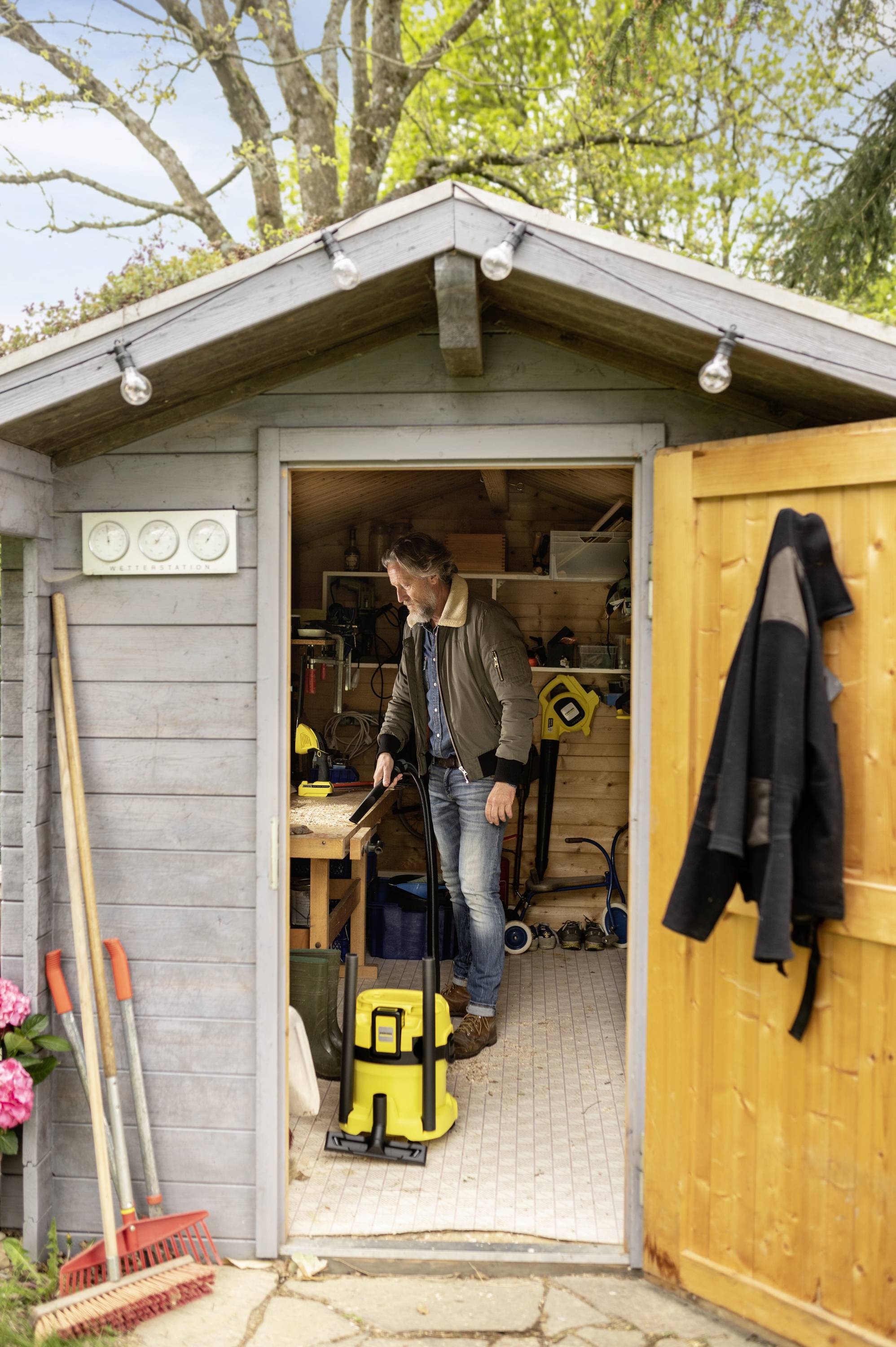 Ein Mann reinigt ein Gartenhaus mit einem gelben Staubsauger. Geöffnete Tür, Werkzeuge und Pflanzen sind sichtbar. Holzhaus im Garten.