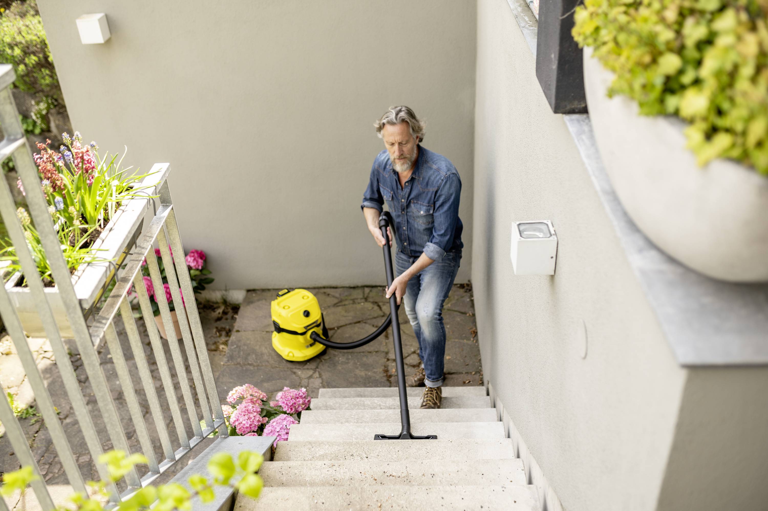 Ein Mann reinigt eine Treppe mit einem gelben Staubsauger im Gartenbereich. Blumen und Pflanzen befinden sich im Hintergrund.