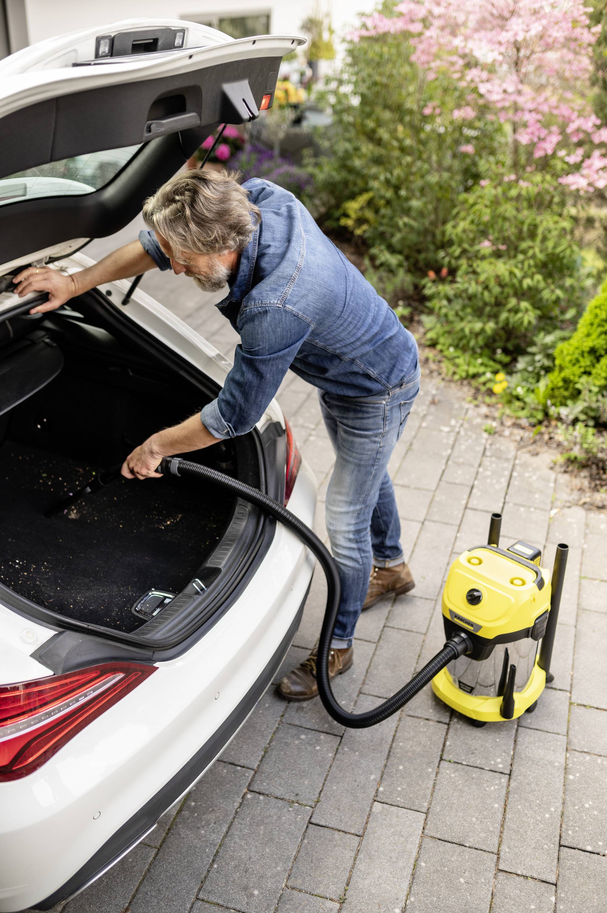 Ein Mann saugt den Kofferraum eines weißen Autos mit einem gelben Staubsauger aus. Er steht auf einer gepflasterten Einfahrt, umgeben von Pflanzen.