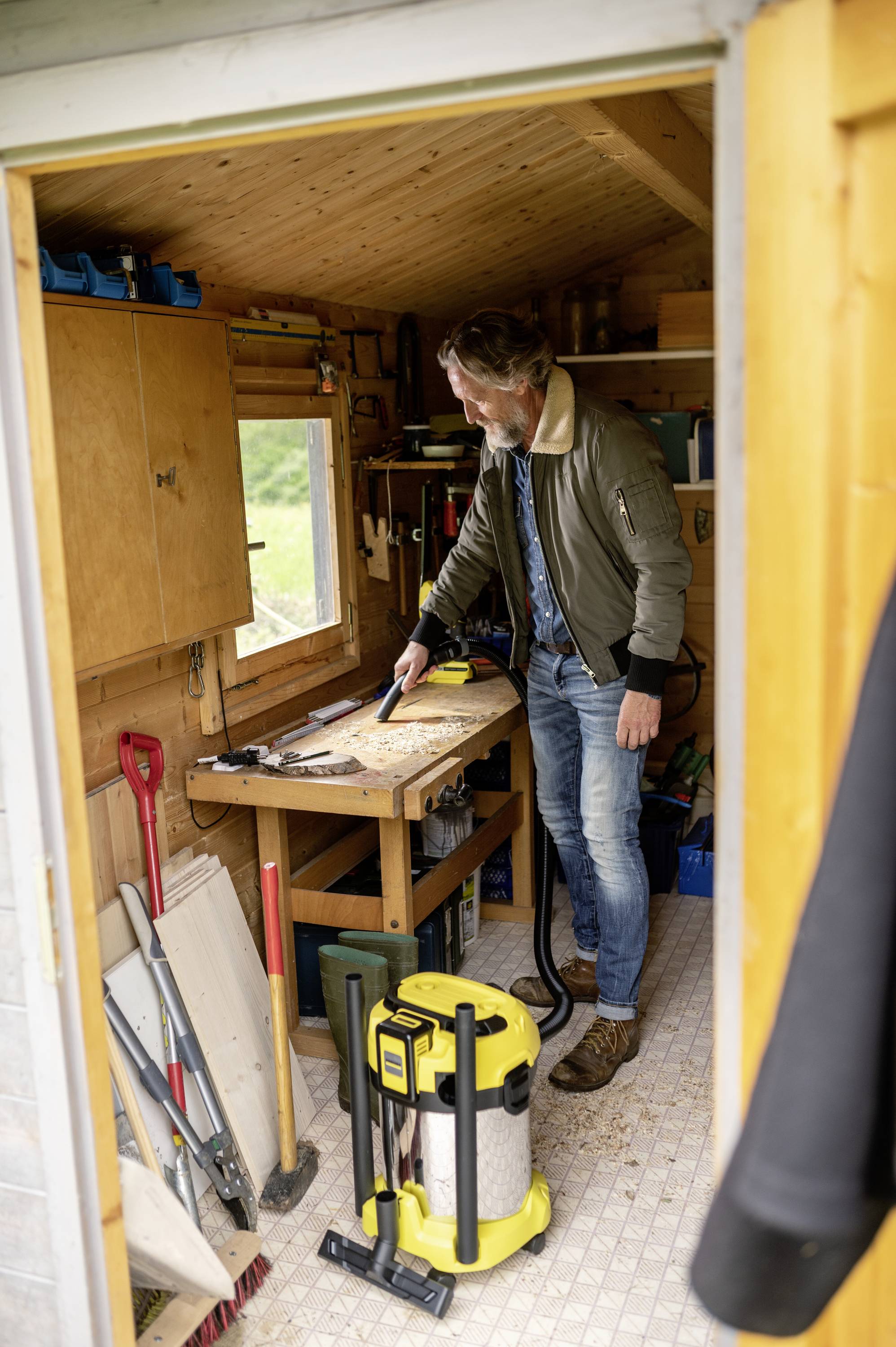 Ein Mann arbeitet an einem Holztisch in einer gut ausgestatteten Werkstatt. Werkzeuge und Materialien sind im Raum verteilt.
