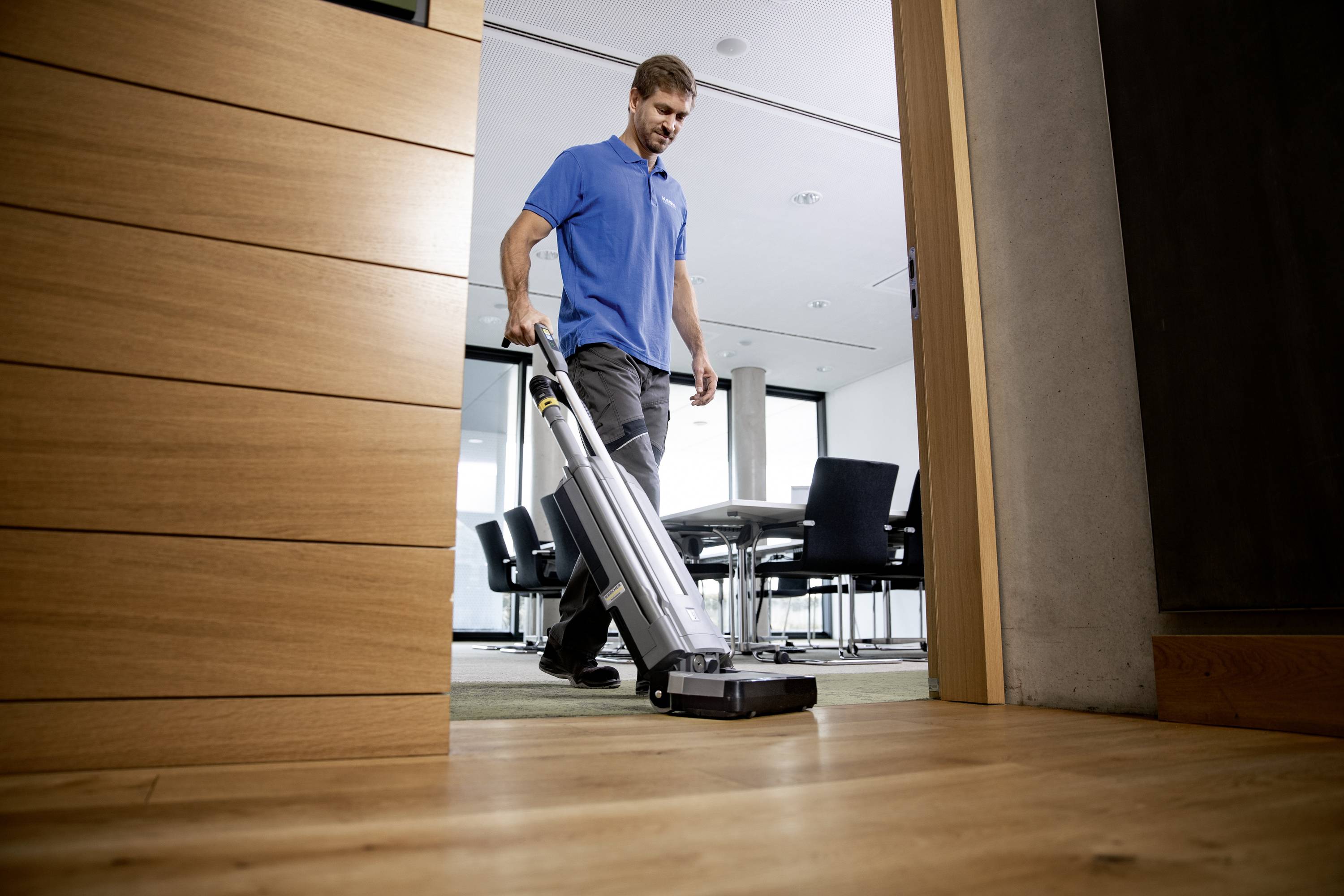 Eine Person saugt mit einem Staubsauger den Boden eines modernen Büros. Im Hintergrund sind Konferenztisch und Stühle sichtbar.