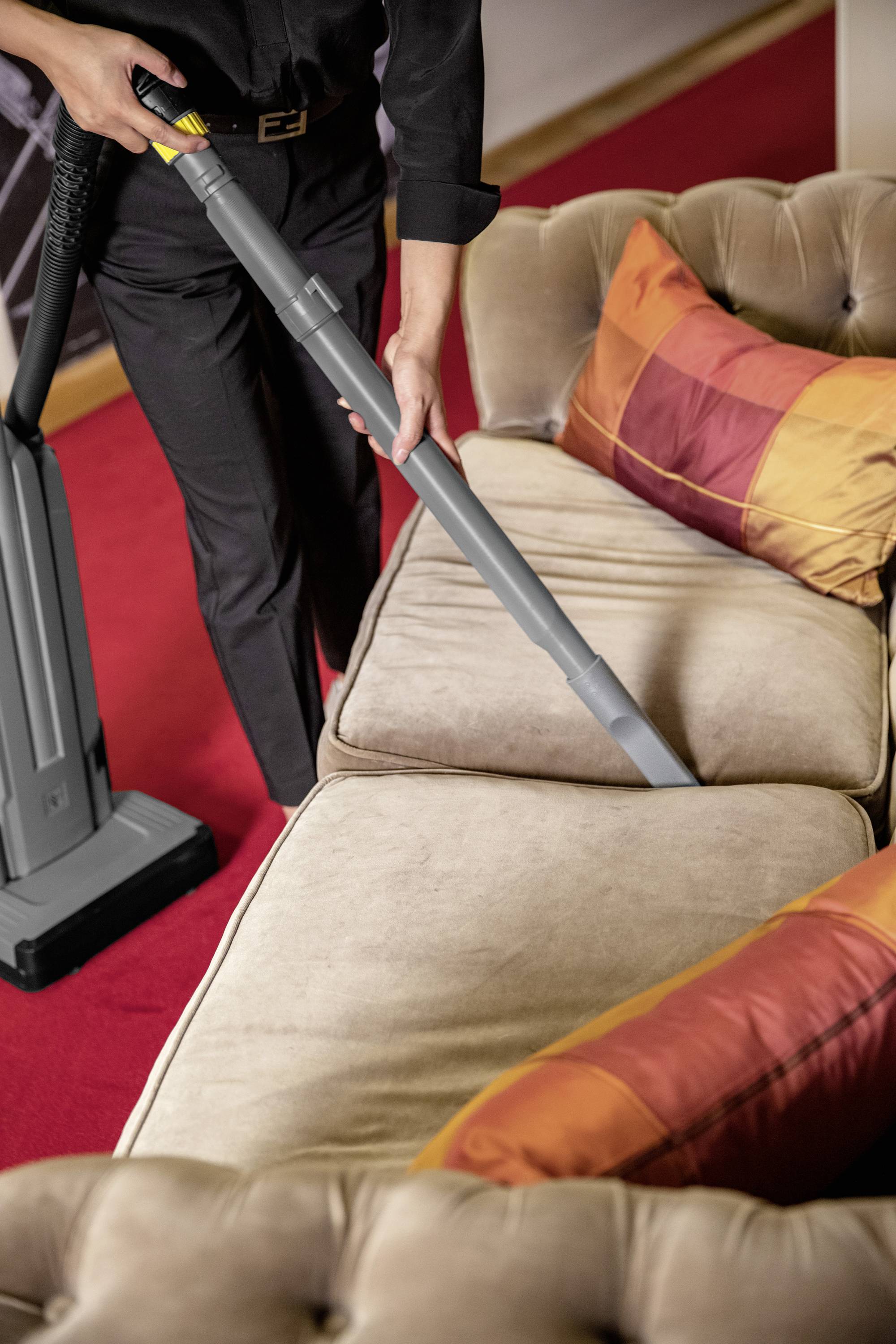 Eine Person saugt ein beigefarbenes Sofa mit einem Staubsauger ab, dabei sind orange und rot karierte Kissen sichtbar.