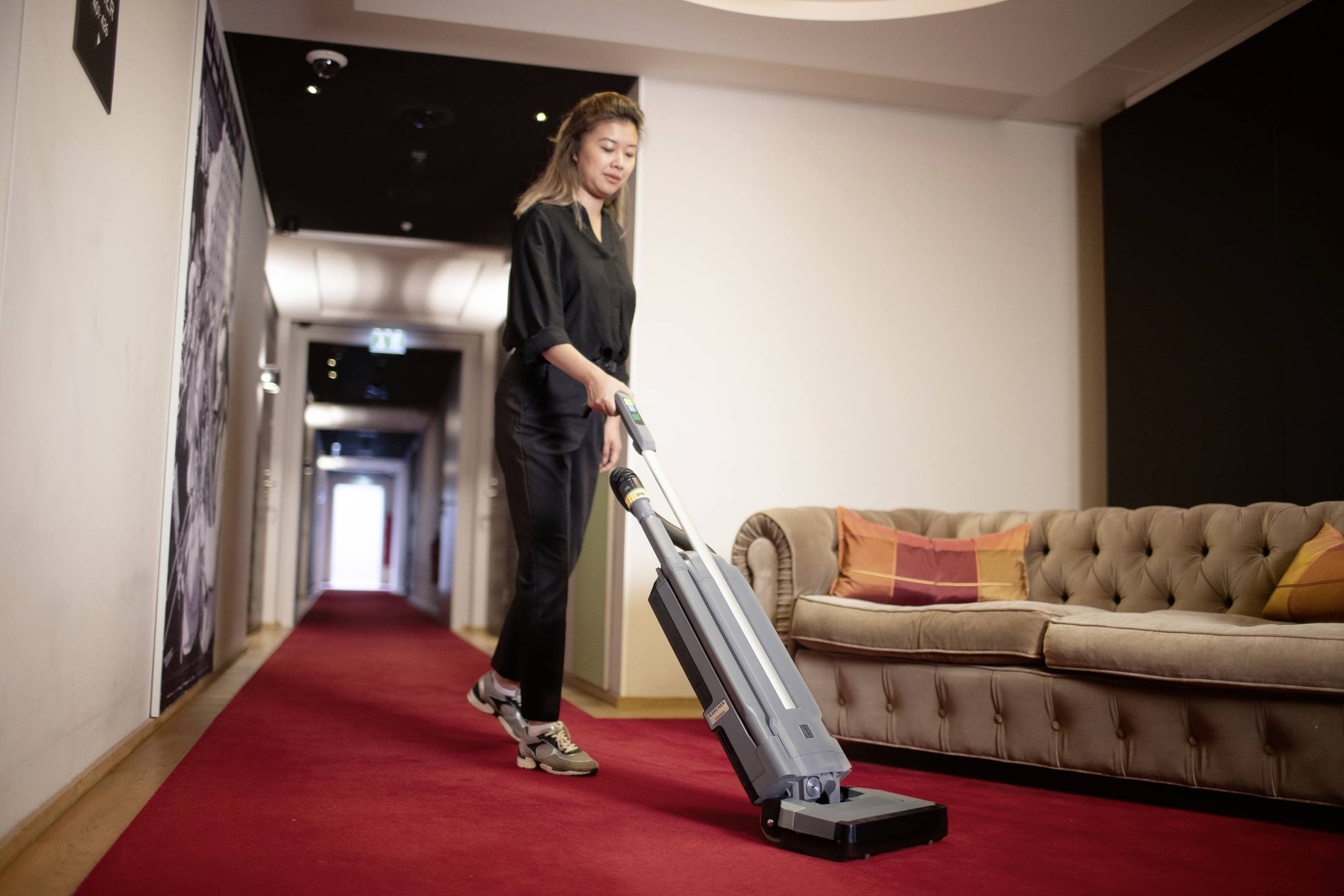 Frau saugt einen roten Teppich in einem Hotelkorridor. Rechts steht ein beige Sofa mit bunten Kissen.