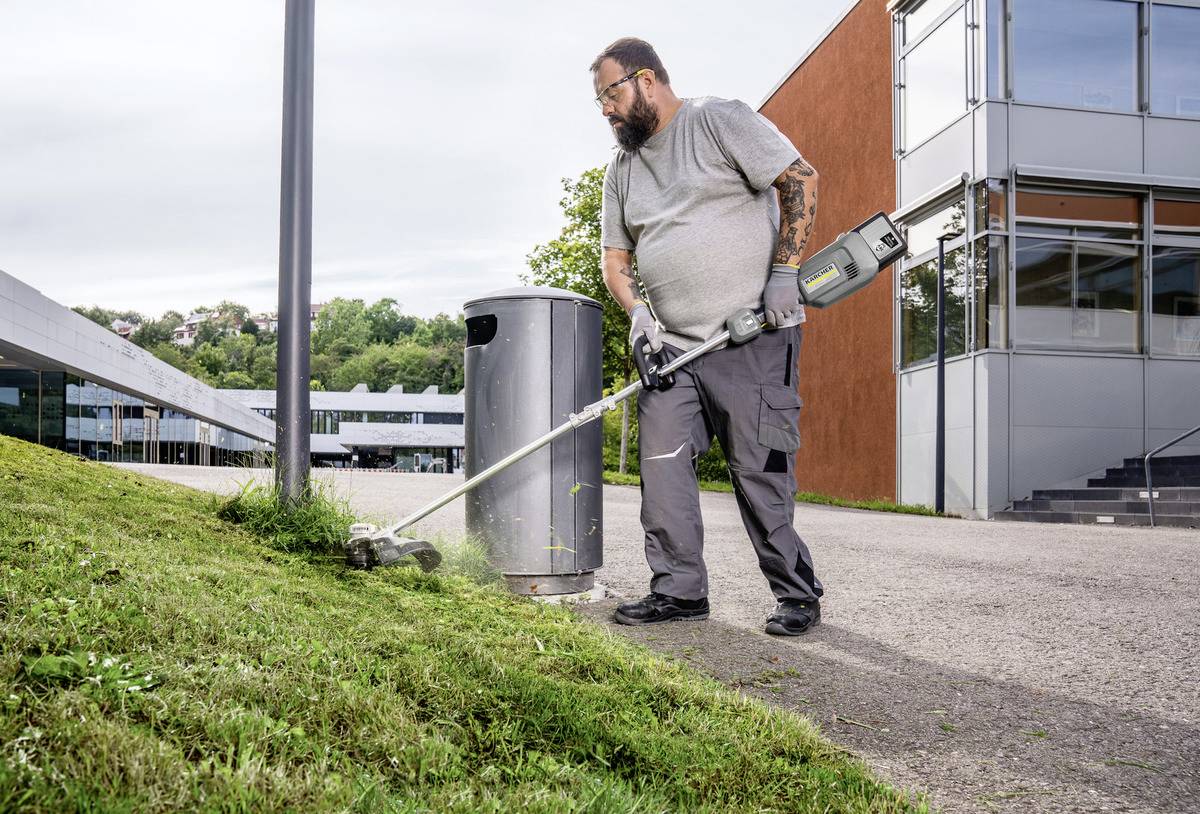 Ein Mann trimmt Gras mit einem Motorsensen in der Nähe eines modernen Gebäudes an einem klaren Tag und konzentriert sich dabei auf den Rasenrand entlang eines Weges und einer Mülltonne.