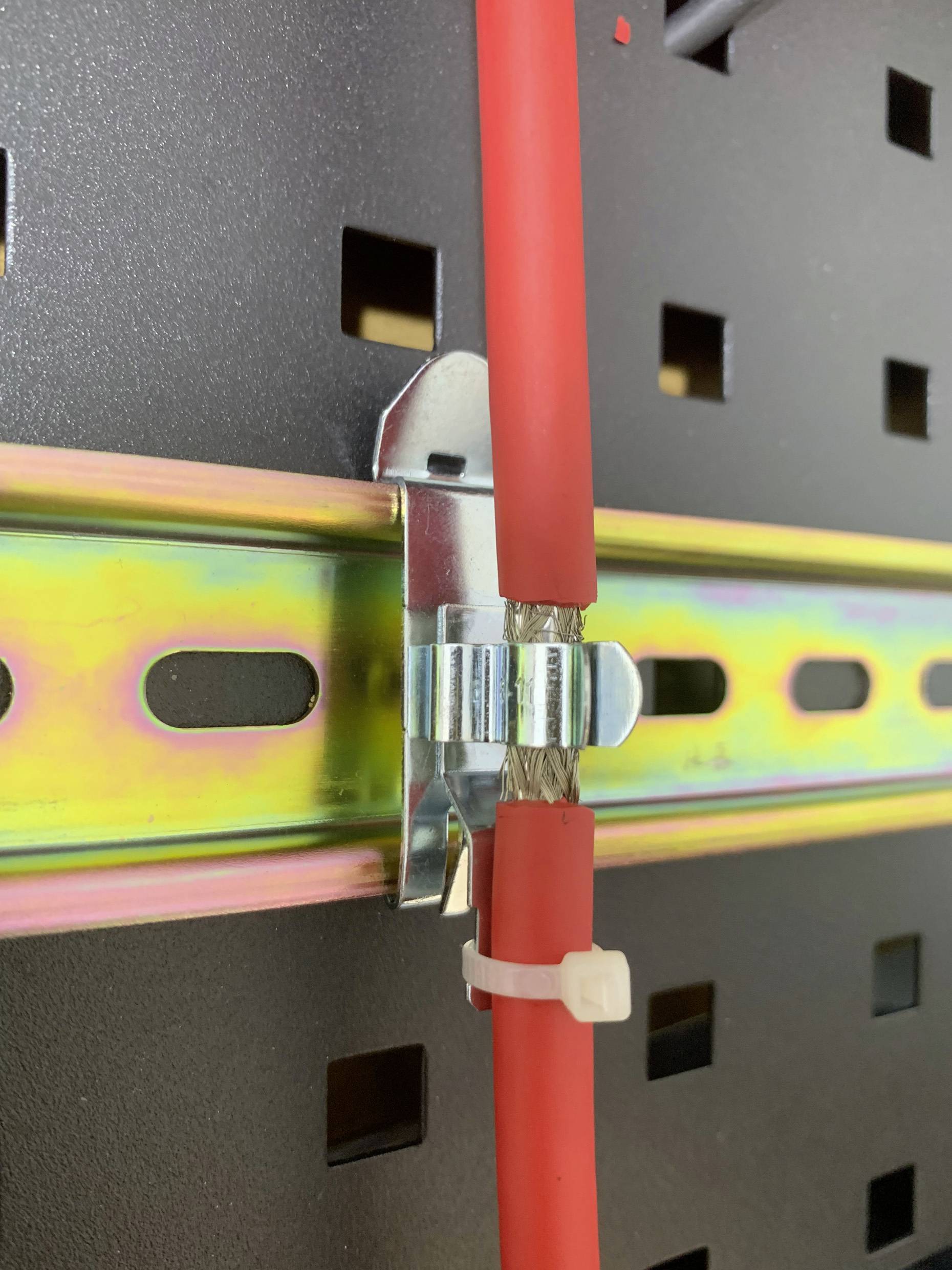Ein rotes Kabel mit Metallschutz ist mit einer Kabelschelle an einer Lochblechwand befestigt, wobei es an einer Regenbogenschiene entlangläuft.