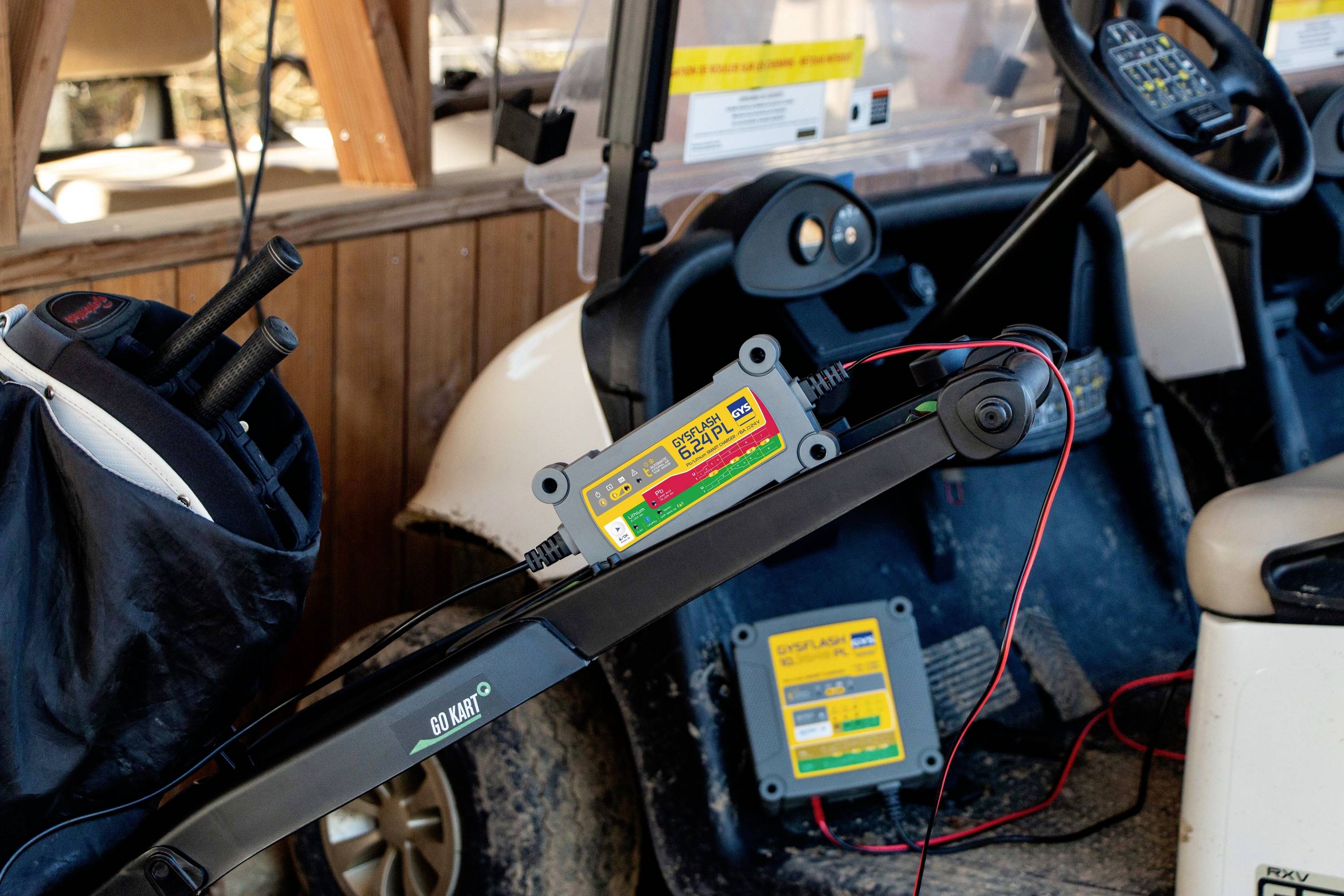 Ein elektrisches Golfcart wird mit einem Ladegerät verbunden. Im Hintergrund sind Golftaschen und -schläger sichtbar.