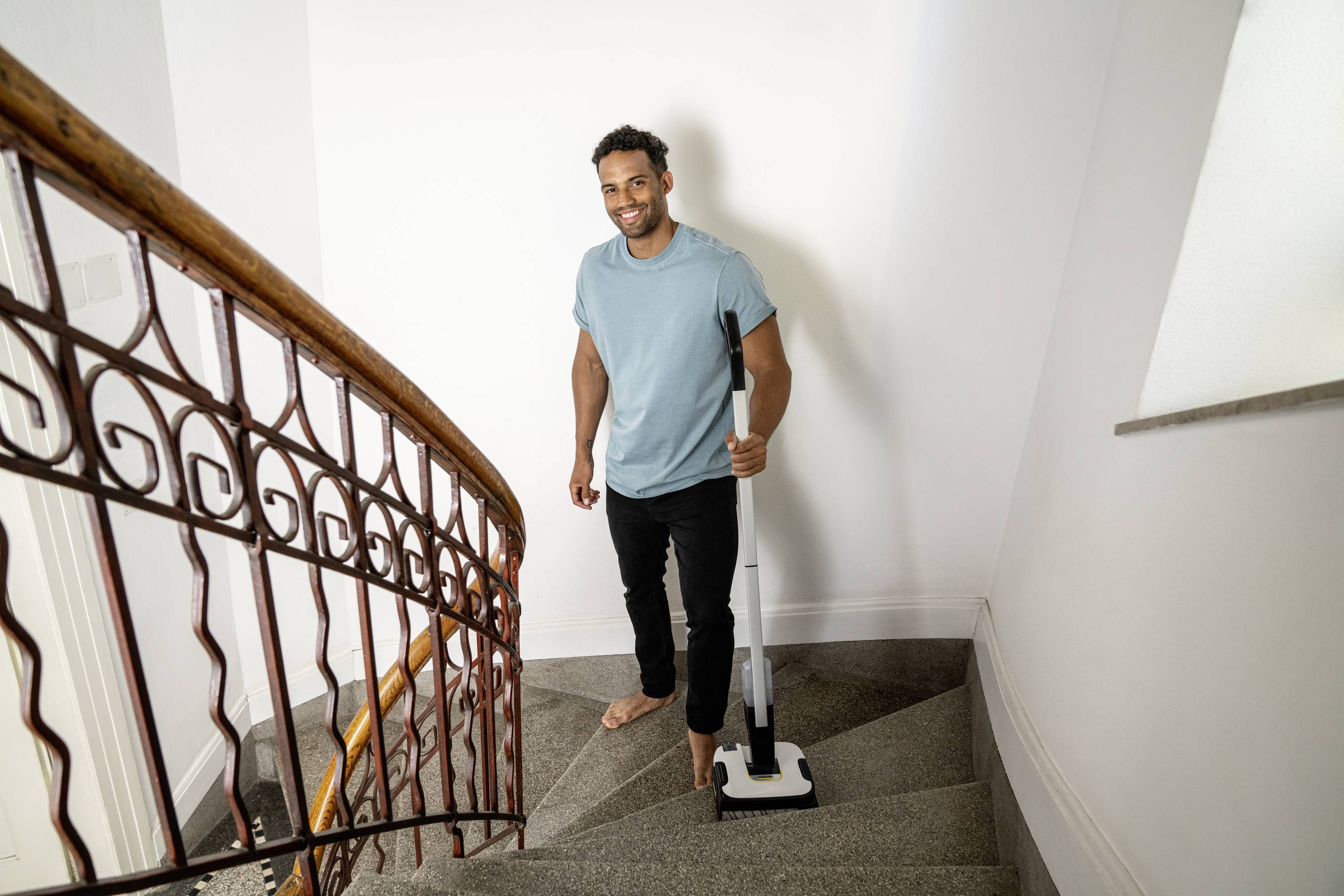 Ein Mann steht barfuß auf einer Treppe und hält einen Staubsauger. Er lächelt und scheint beim Reinigen zu sein.