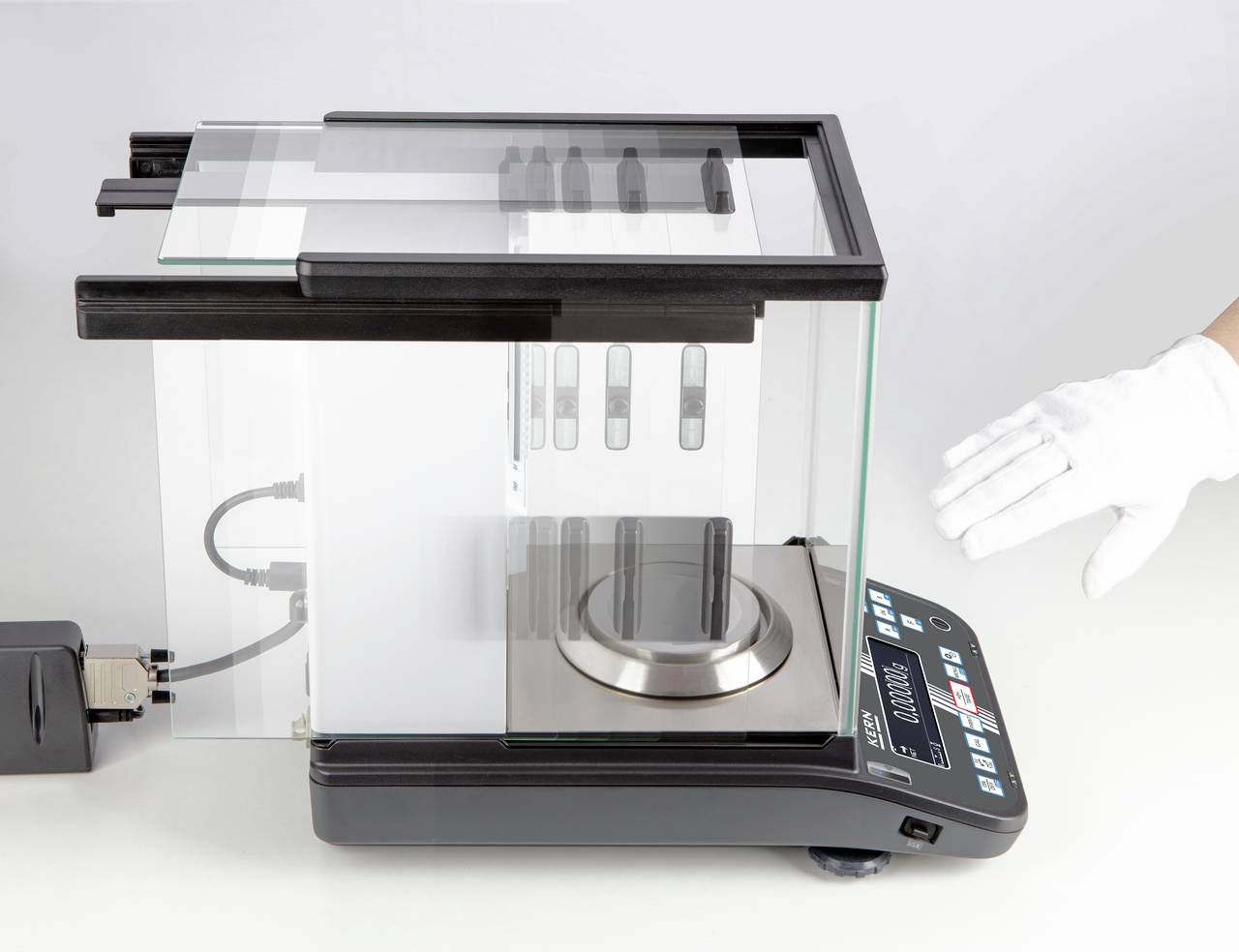 Elektronische Präzisionswaage mit Glasgehäuse, Display und Steuerungstasten. Hand mit Handschuh zeigt auf die Waage.