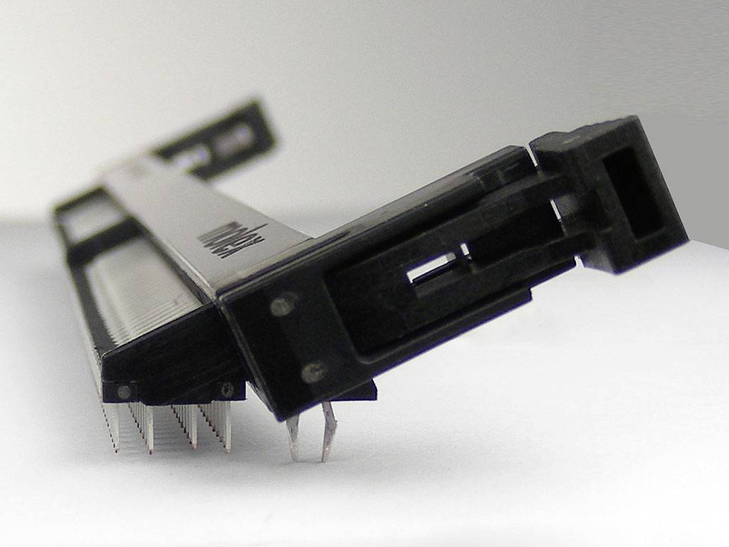 Ein schwarzer Schiebepotentiometer mit mehreren Anschlüssen auf einer weißen Oberfläche, leicht schräg von der Seite aufgenommen.