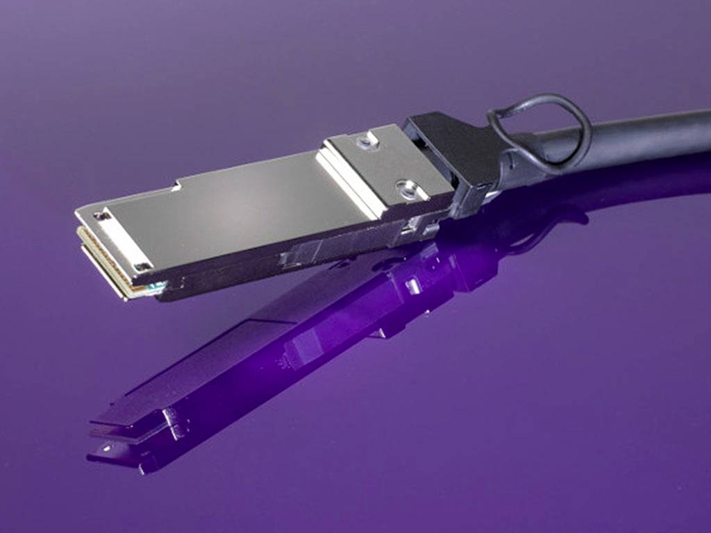 Ein silberner Computer-Stecker liegt auf einer lila Oberfläche, das Kabel zeigt nach rechts. Reflektion des Steckers auf der Oberfläche sichtbar.
