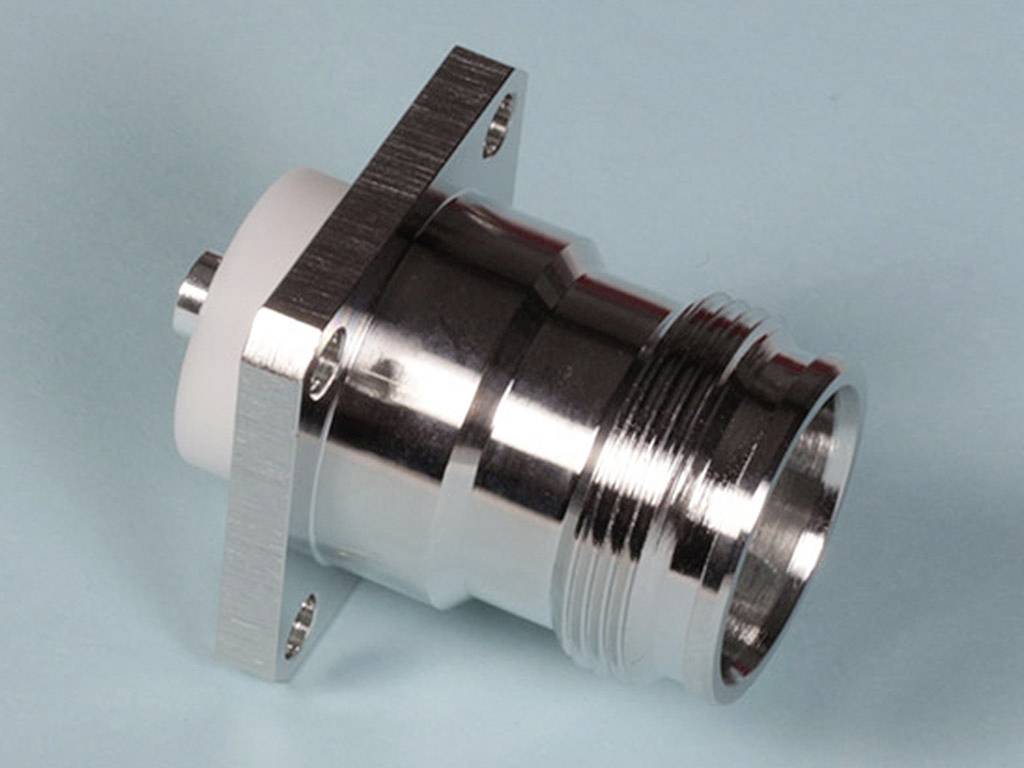 Ein metallischer Koaxialstecker auf hellblauem Hintergrund. Der Stecker hat ein rundes Gewinde und eine rechteckige Basis mit Schraublöchern.