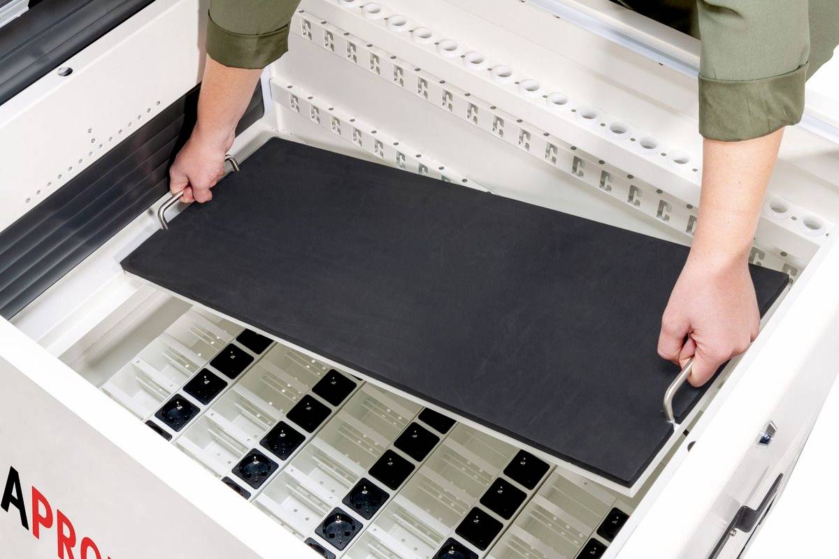 Eine Person hebt ein schwarzes Panel aus einer Lade mit Steckdosen heraus. Die Szene deutet auf eine technische oder elektronische Umgebung hin.