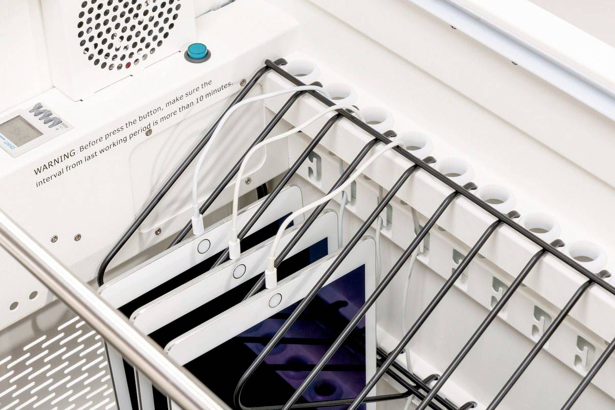 Tablett-Aufbewahrungsstation mit mehreren Tablets, die vertikal in Fächer gesteckt und über Kabel aufgeladen werden. Warnhinweis ist sichtbar.