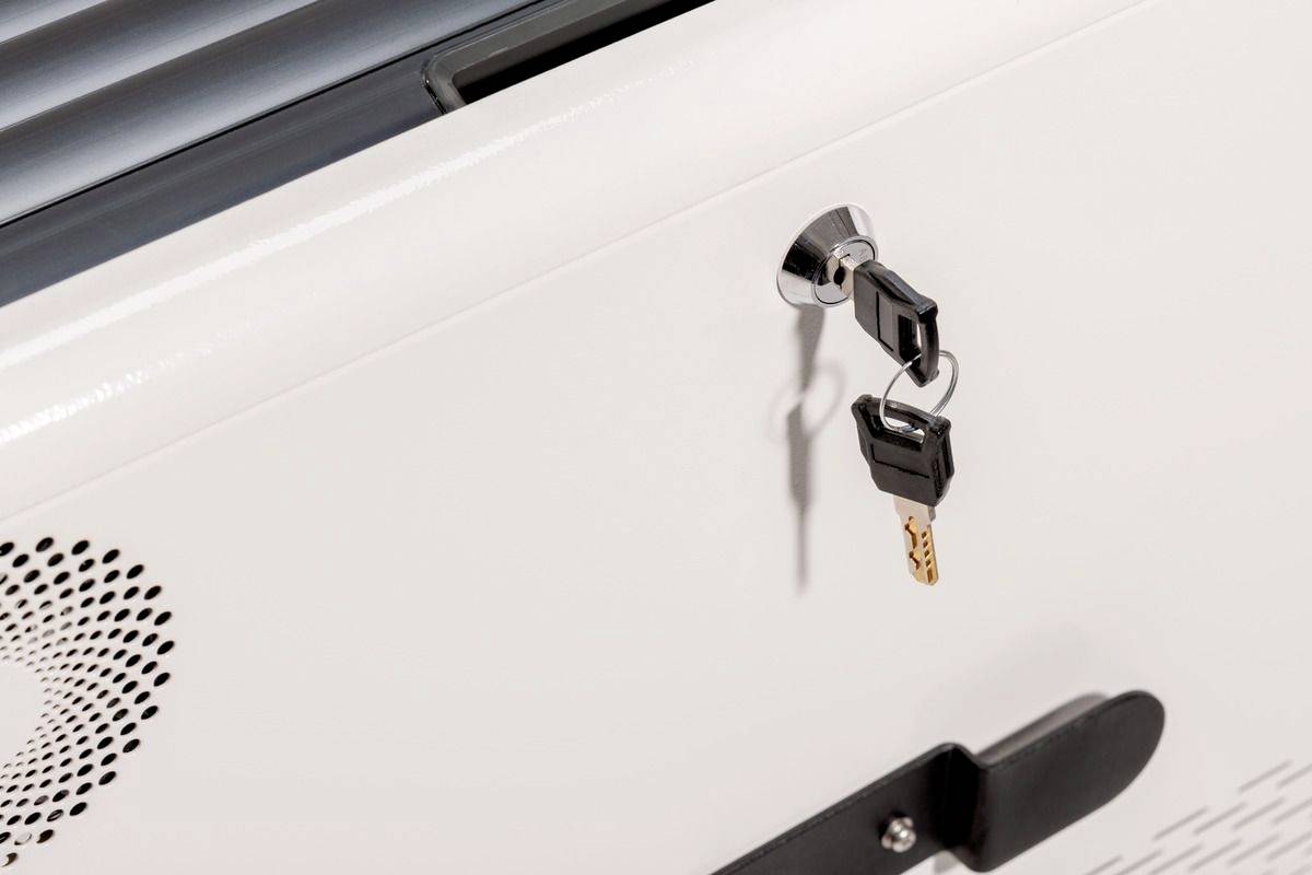 Ein Schlüssel steckt in einem Schloss an einem weißen Metallschrank, daneben ist ein Lüftungsgitter zu sehen.