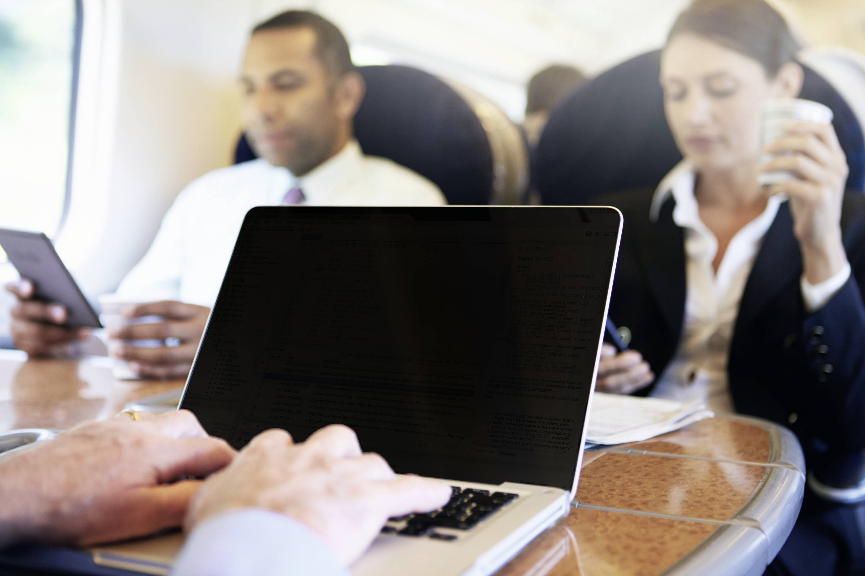 Eine Person arbeitet auf einem Laptop in einem Zug, während zwei weitere Personen im Hintergrund ein Tablet und eine Kaffeetasse halten.