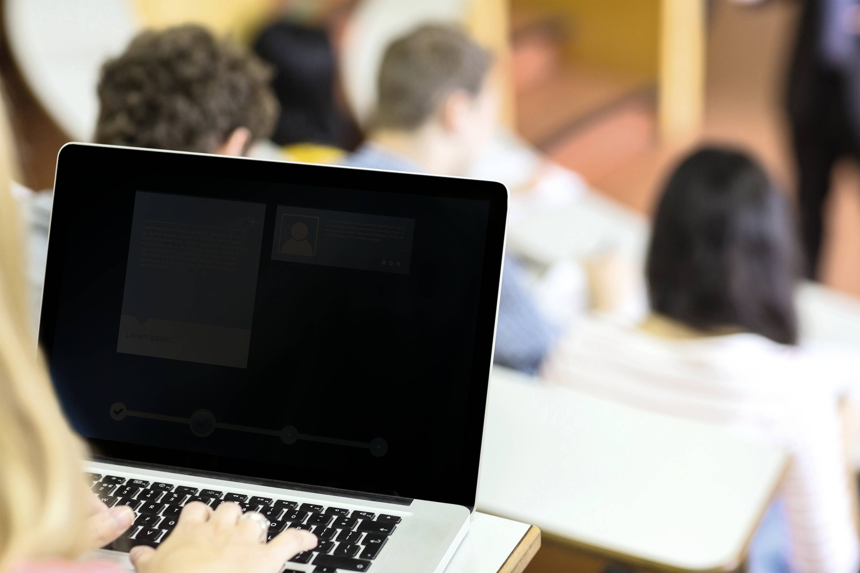 Eine Person arbeitet an einem Laptop in einem Hörsaal, während andere Studierende im Hintergrund einem Vortrag folgen.