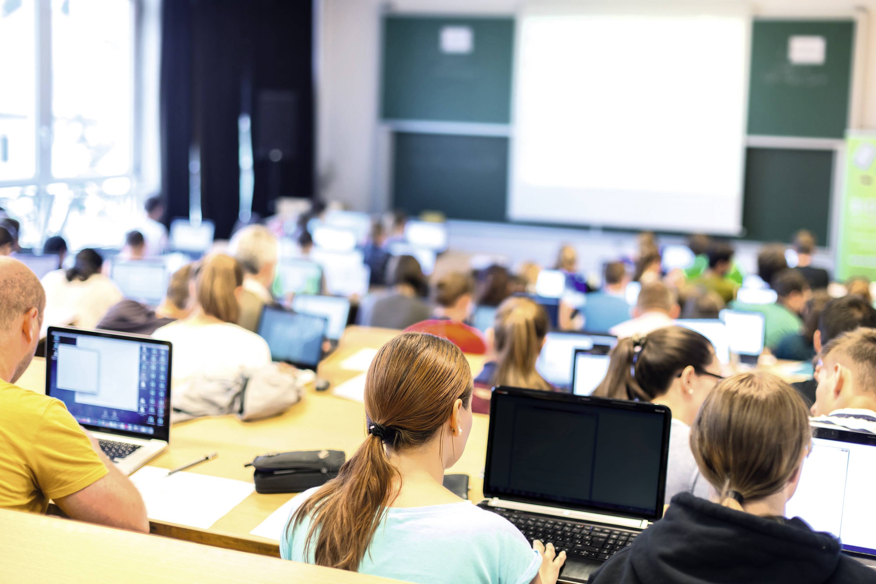 Studierende in einem Klassenzimmer sitzen an Tischen und arbeiten auf Laptops. Im Hintergrund sind Tafeln und ein Projektor sichtbar.