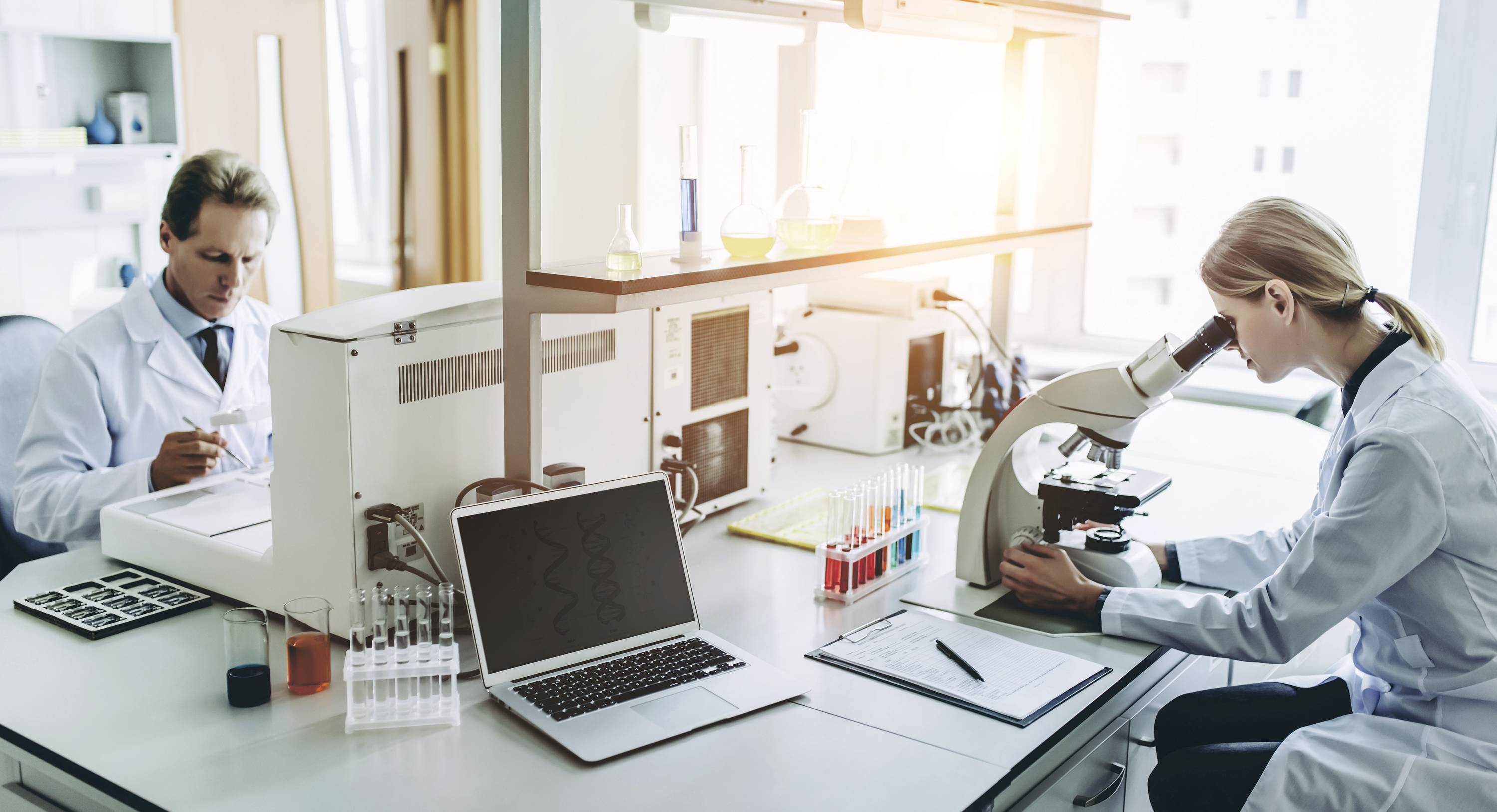 Zwei Wissenschaftler arbeiten in einem Labor. Eine Person benutzt ein Mikroskop, die andere notiert Ergebnisse. Verschiedene Laborausrüstungen sind sichtbar.