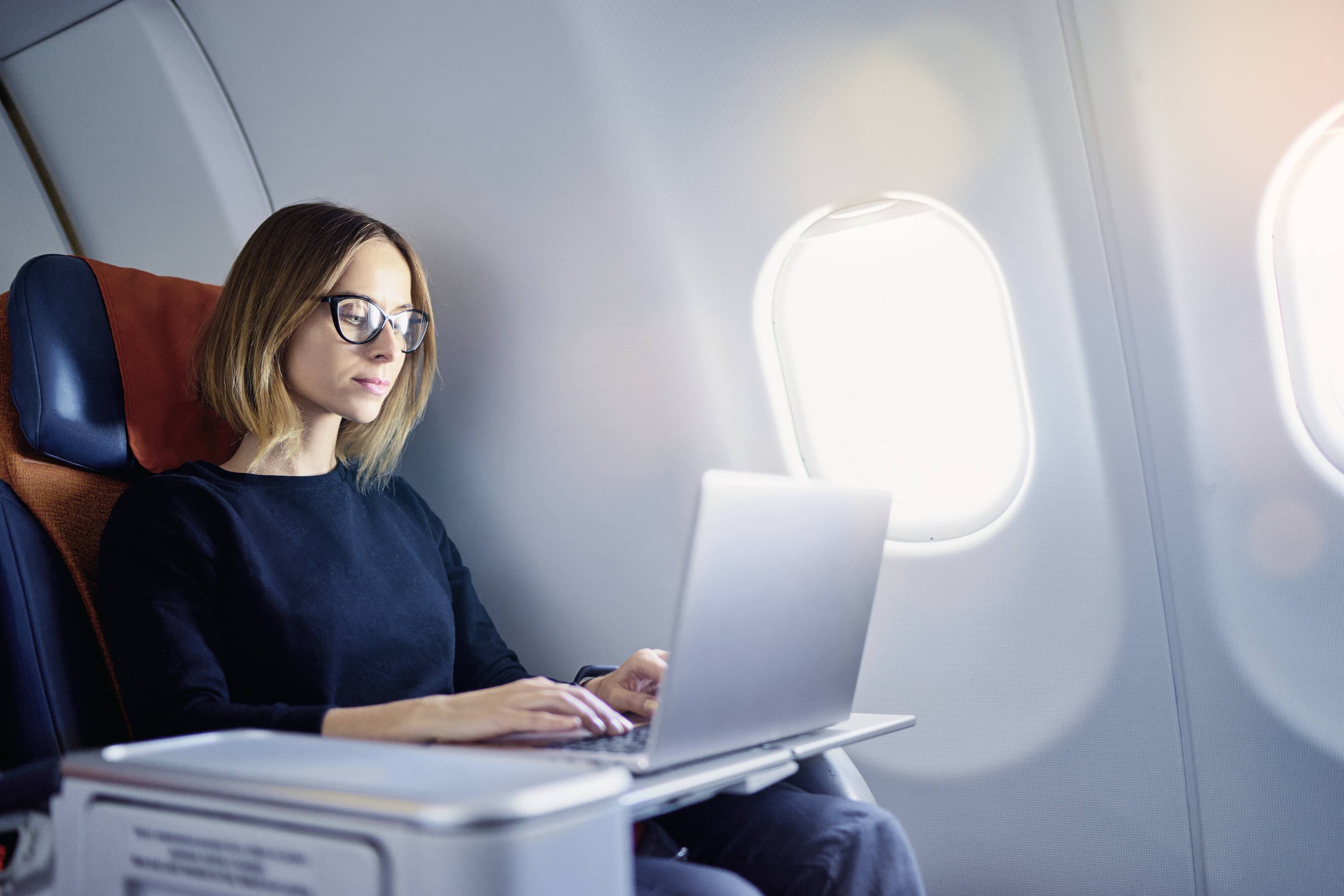 Eine Frau mit Brille arbeitet an einem Laptop, während sie in einem Flugzeug sitzt. Sie ist in einem Sitz am Fenster.