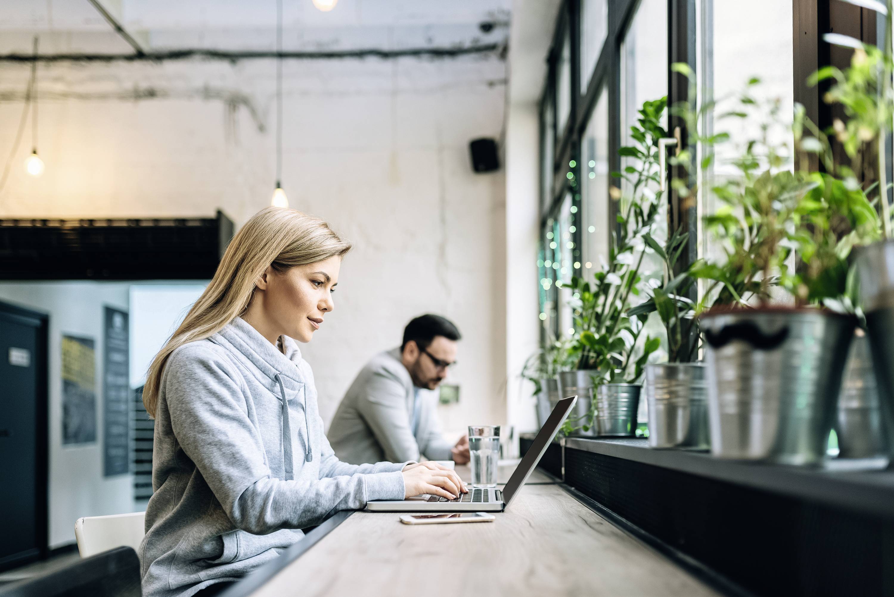 Eine Frau und ein Mann arbeiten an Laptops in einem modernen Café, umgeben von Pflanzen auf der Fensterbank.
