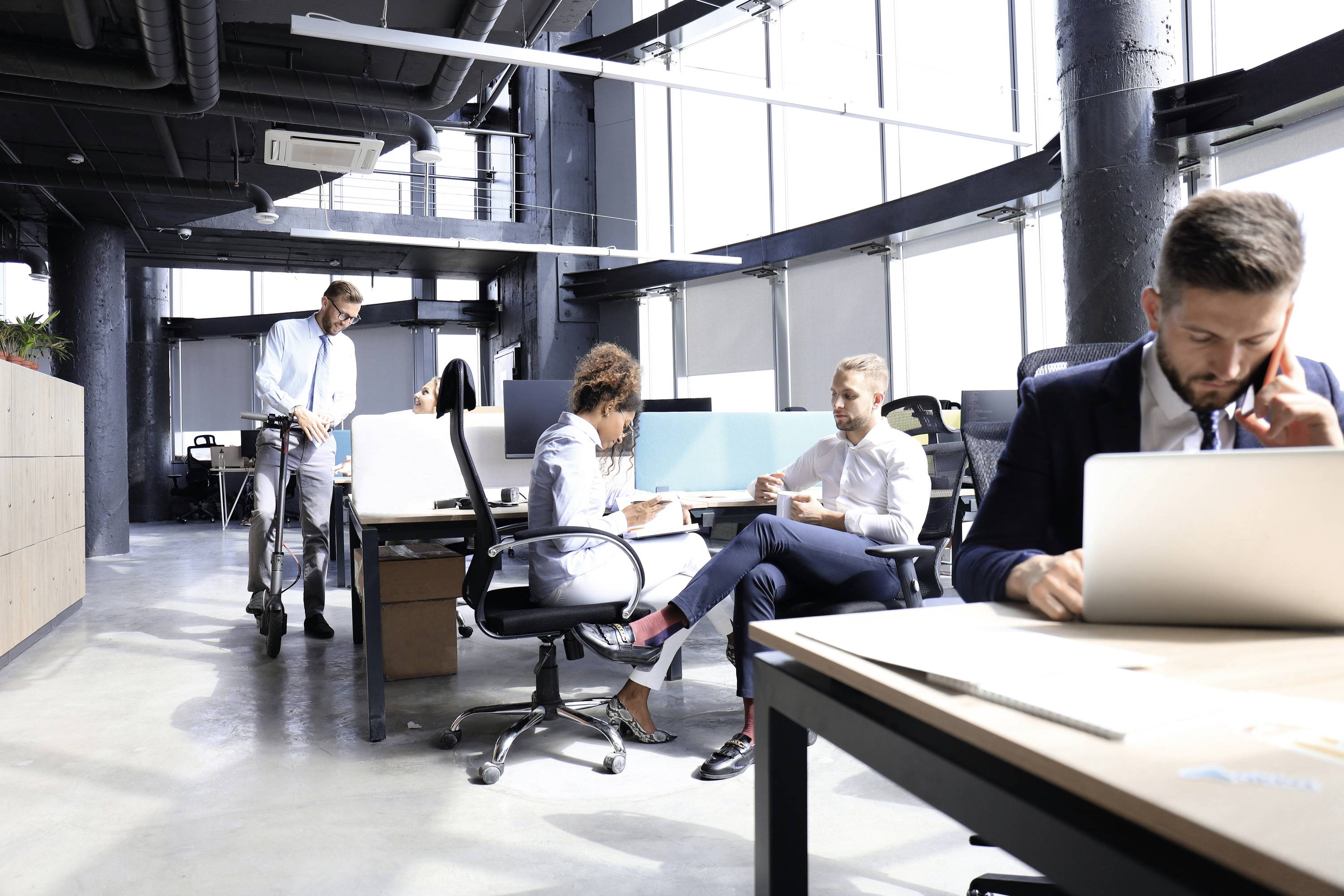 Mehrere Personen arbeiten in einem modernen Büro mit großen Fenstern. Eine Person arbeitet am Laptop, andere diskutieren an einem Tisch.