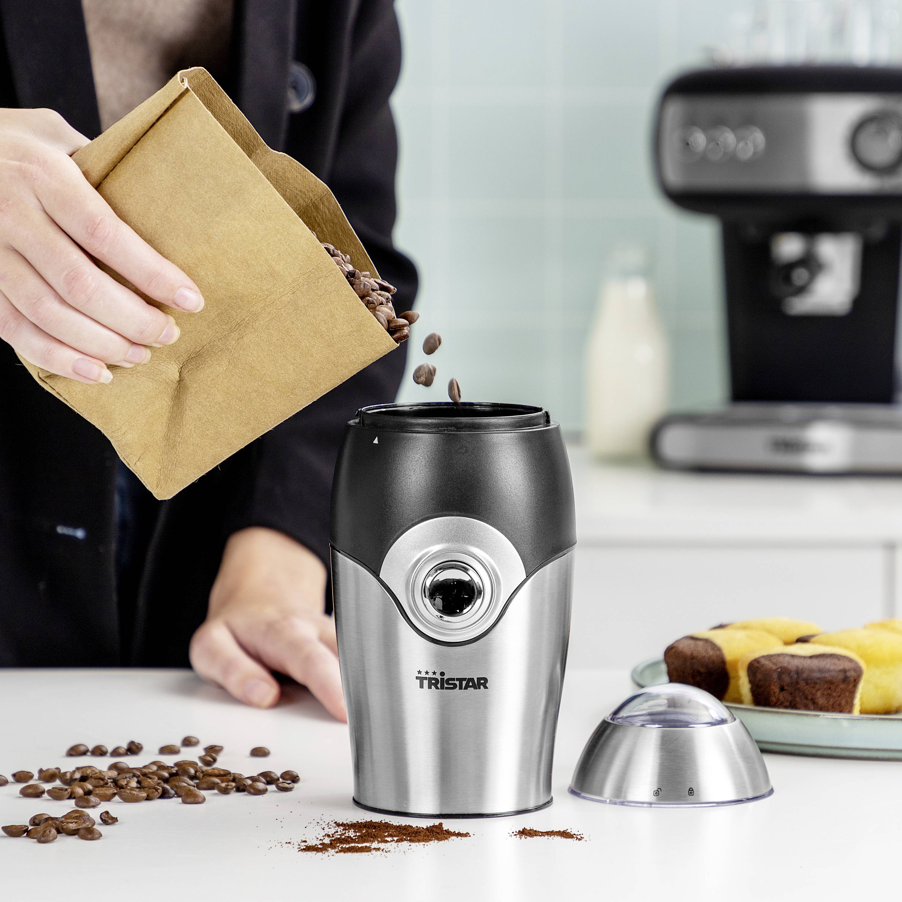 Eine Person füllt Bohnen in eine silberne Kaffeemühle auf einem Küchentresen. Daneben liegen Kaffeebohnen und Muffins.