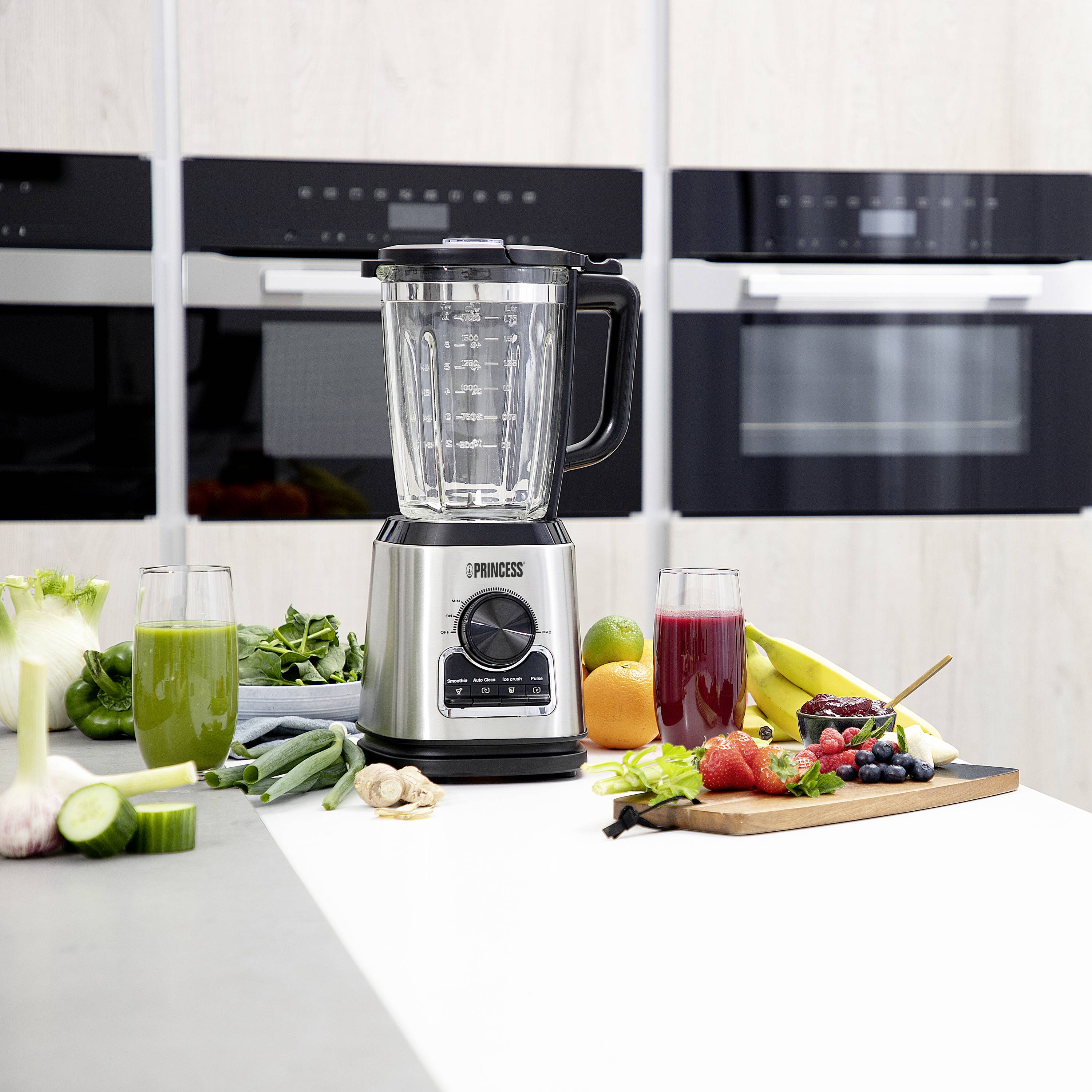 Ein Mixer auf einer Küchentheke, umgeben von frischem Gemüse, Obst und zwei Gläsern Smoothies in Grün und Rot. Im Hintergrund Küchengeräte.