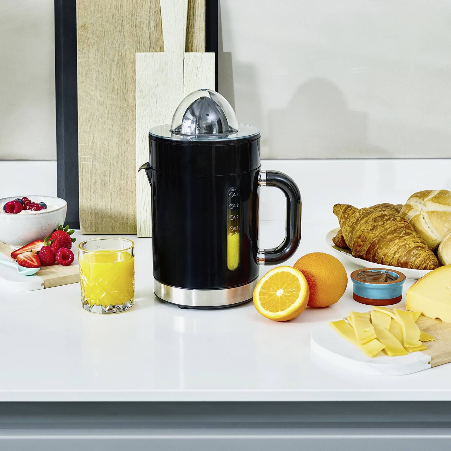 Ein Frühstückstisch mit einer elektrischen Saftpresse, frischem Orangensaft, Croissants, Brötchen, Orangen, Marmelade, Käse und Beeren.