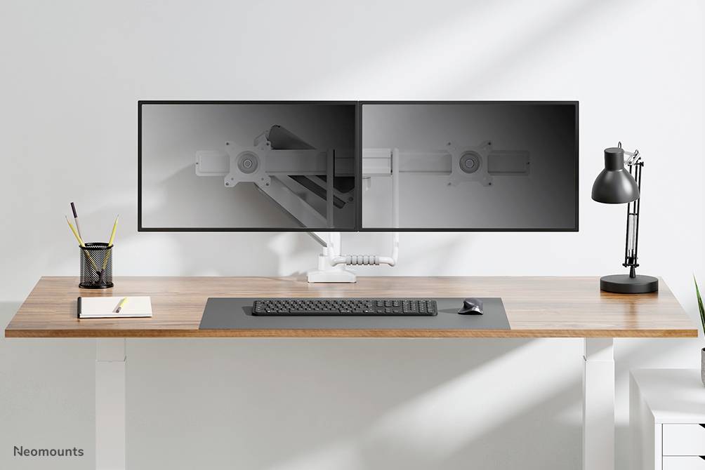 Ein Holztisch mit Tastatur, Maus, zwei nebeneinandergestellten Monitoren und einer schwarzen Schreibtischlampe. Links steht eine Tasse.