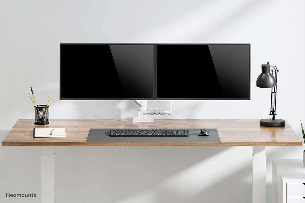 Ein aufgeräumter Schreibtisch mit zwei nebeneinanderliegenden Monitoren, einer Tastatur, Lampe, Notizbuch und Stiftbehälter.