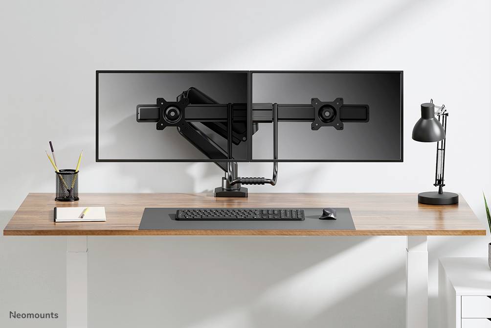 Zwei leere Monitorhalterungen auf einem Schreibtisch mit einer Lampe, Tastatur und Notebook. Schlichtes, modernes Bürodesign.